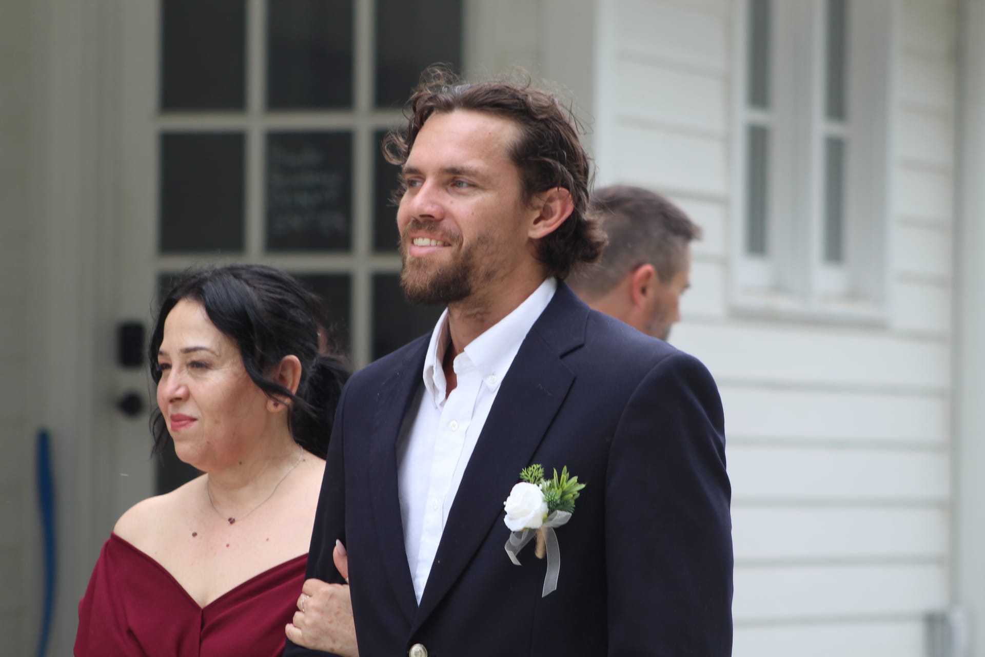 Smiling groom in a navy suit with a white rose boutonniere escorts a bridesmaid in a burgundy off-shoulder dress outside a historic white manor wedding venue.