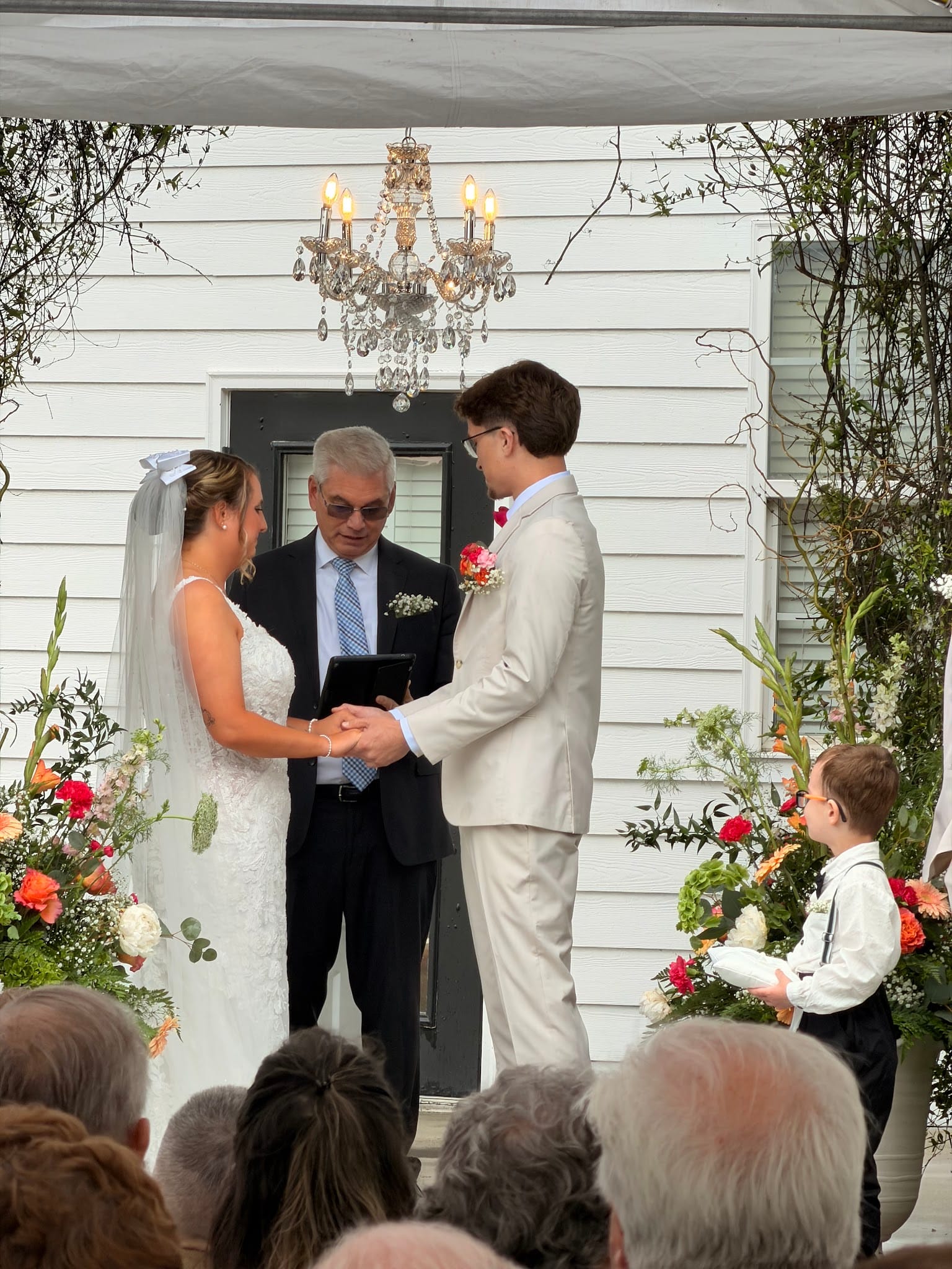 Bride and groom exchange vows outdoors at Scarborough House estate, surrounded by lush coral and white floral arrangements beneath a crystal chandelier, with an officiant and young ring bearer beside them.