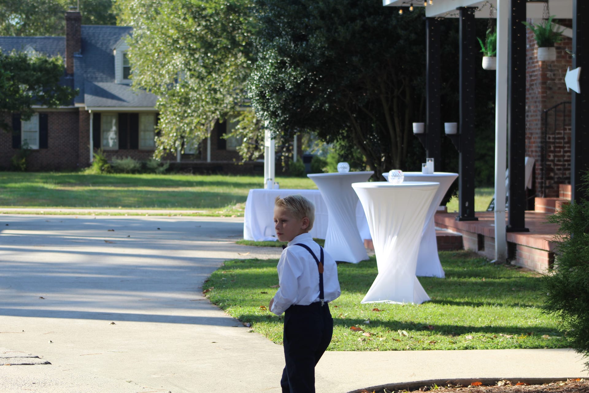 A young boy in a white dress shirt and navy suspenders stands on the pathway during cocktail hour at Scarborough House historic estate wedding venue, with draped cocktail tables and lush trees in the background.