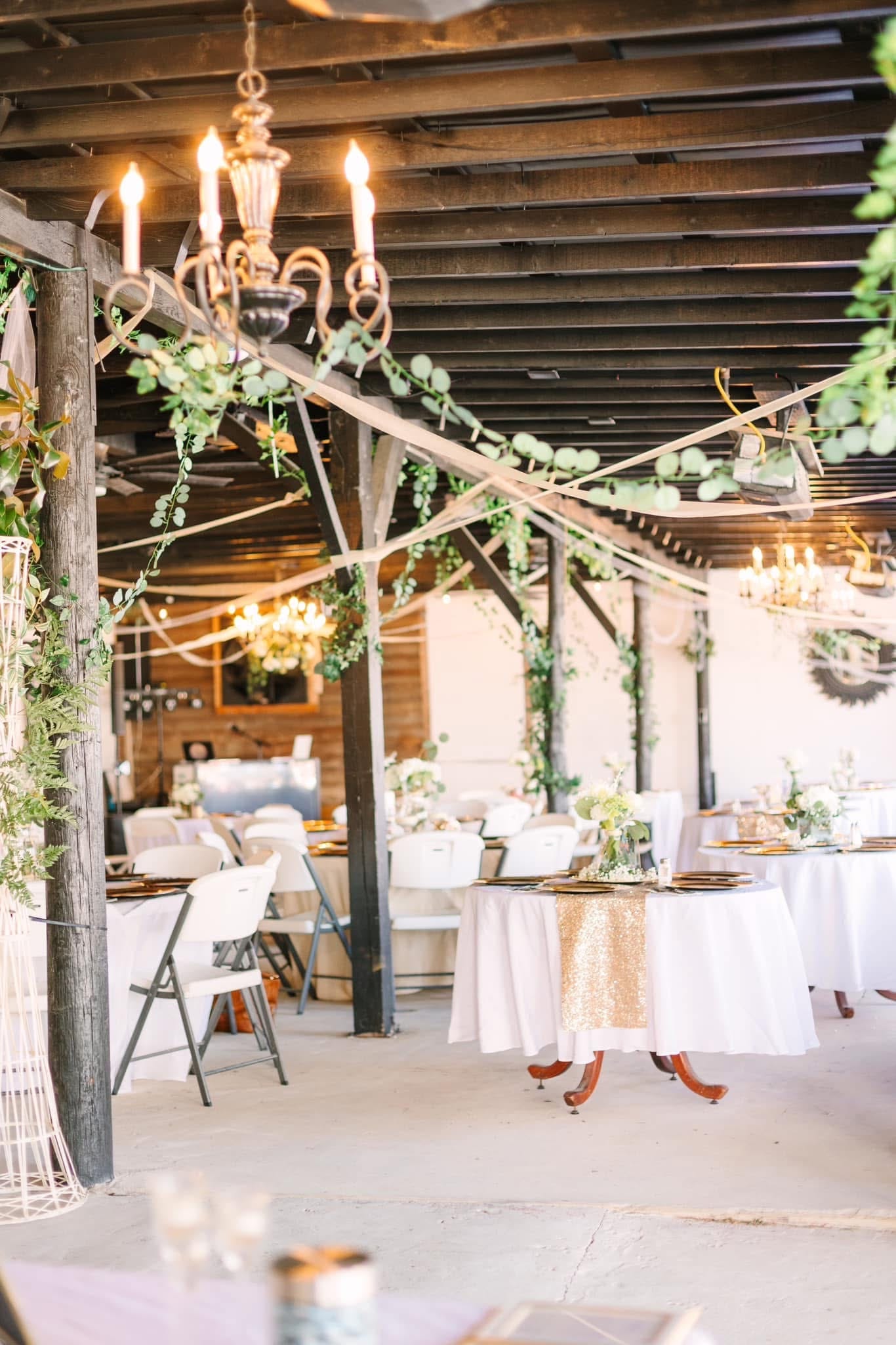 Elegantly decorated wedding reception space featuring candlelit chandeliers draped with eucalyptus garlands, white linen tables with gold sequin runners, and rustic wooden beam ceilings at Scarborough House.