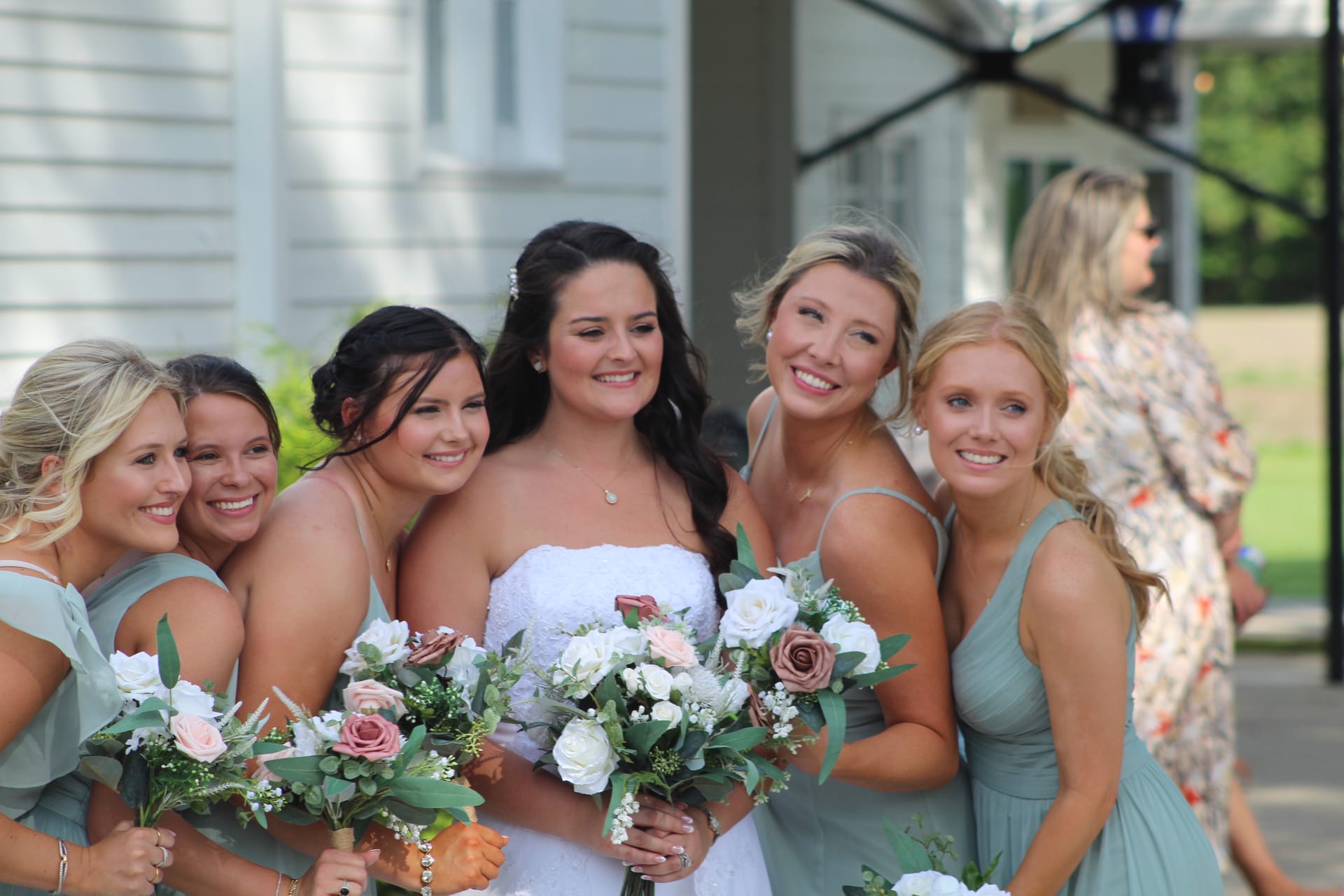 Bride in a strapless white gown laughing with four bridesmaids in sage green dresses holding bouquets of white and blush roses outside a historic manor house.