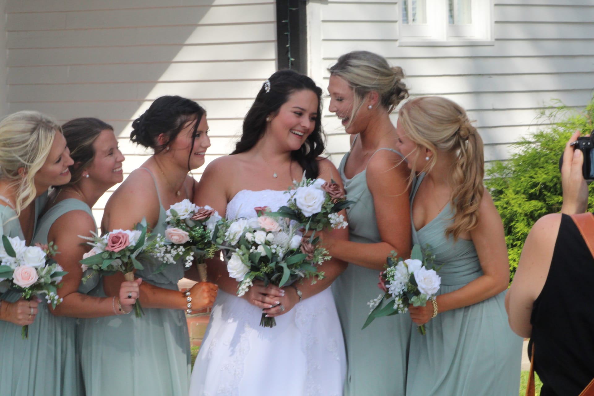 A radiant bride in white holding a lush floral bouquet shares a joyful laughing moment with five bridesmaids in sage green gowns in front of a historic white manor house.