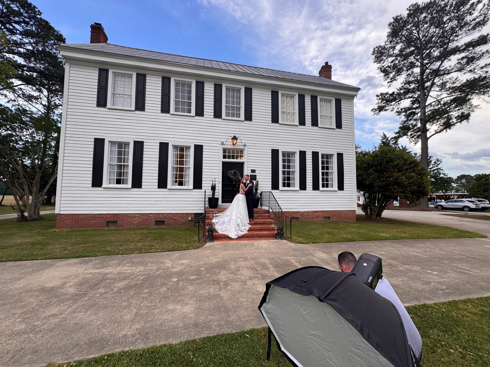 A bride and groom share a romantic embrace on the brick front steps of the 1821 Scarborough House manor while a photographer with a large softbox captures their portrait.