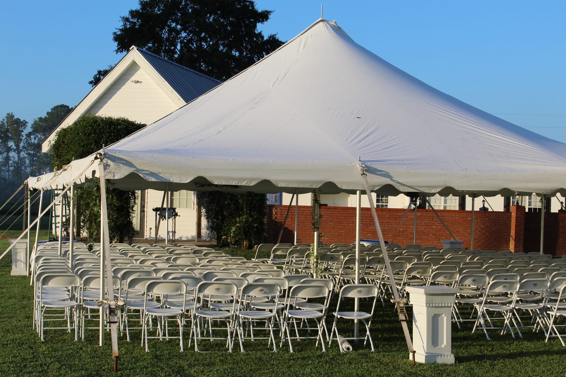 Rows of white folding chairs arranged under a large white pole tent on the ceremony lawn at Scarborough House with the historic white manor and brick wall visible in the background.