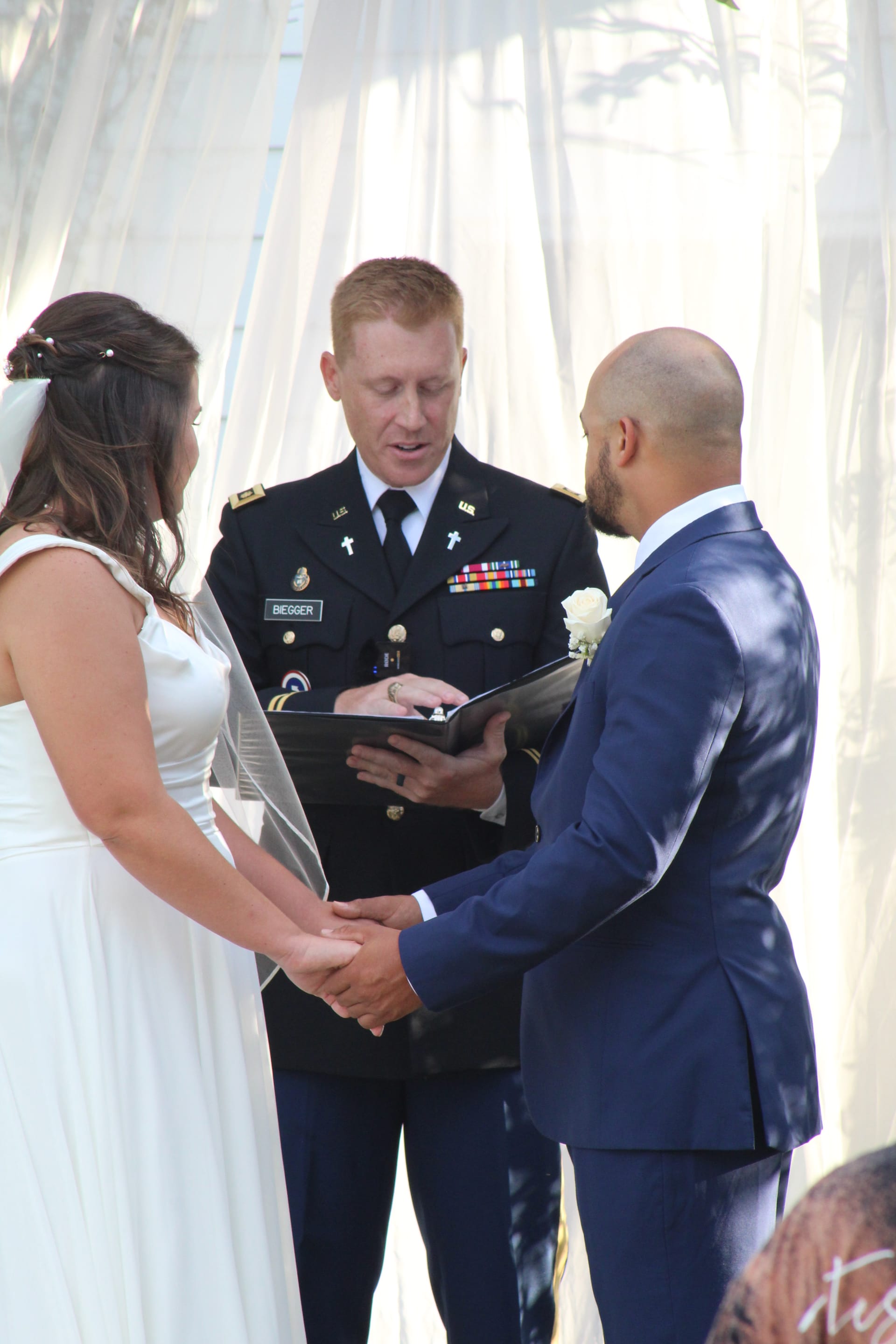 Bride in white gown and groom in navy suit hold hands during their outdoor wedding ceremony officiated by a US Army chaplain beneath white draping.
