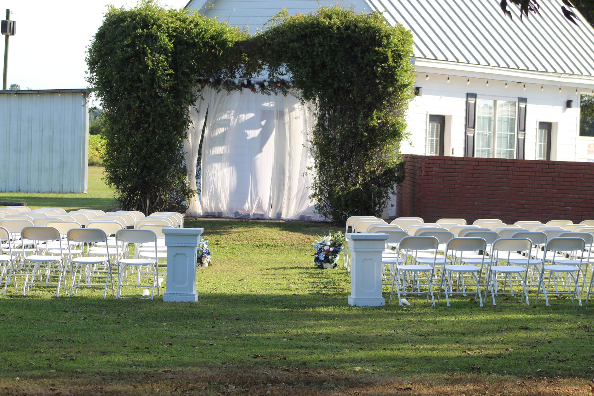 Outdoor wedding ceremony setup at Scarborough House featuring rows of white folding chairs flanking a central aisle leading to a lush ivy-covered arch draped in white fabric and adorned with floral arrangements on a sunny day.
