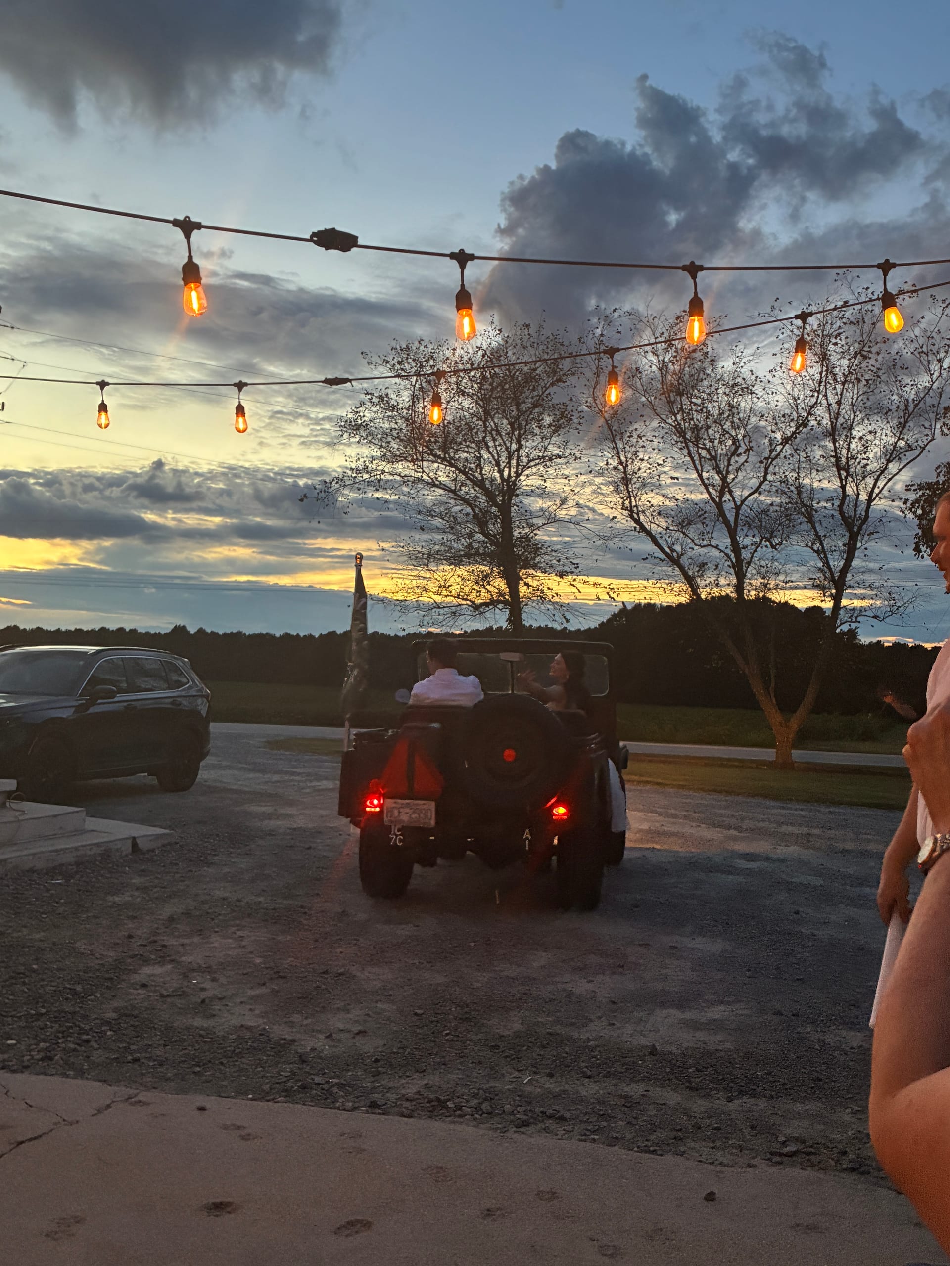 Newlyweds depart in a classic open-top vehicle beneath glowing Edison string lights at dusk with a stunning golden sunset over the farmland fields at Scarborough House.