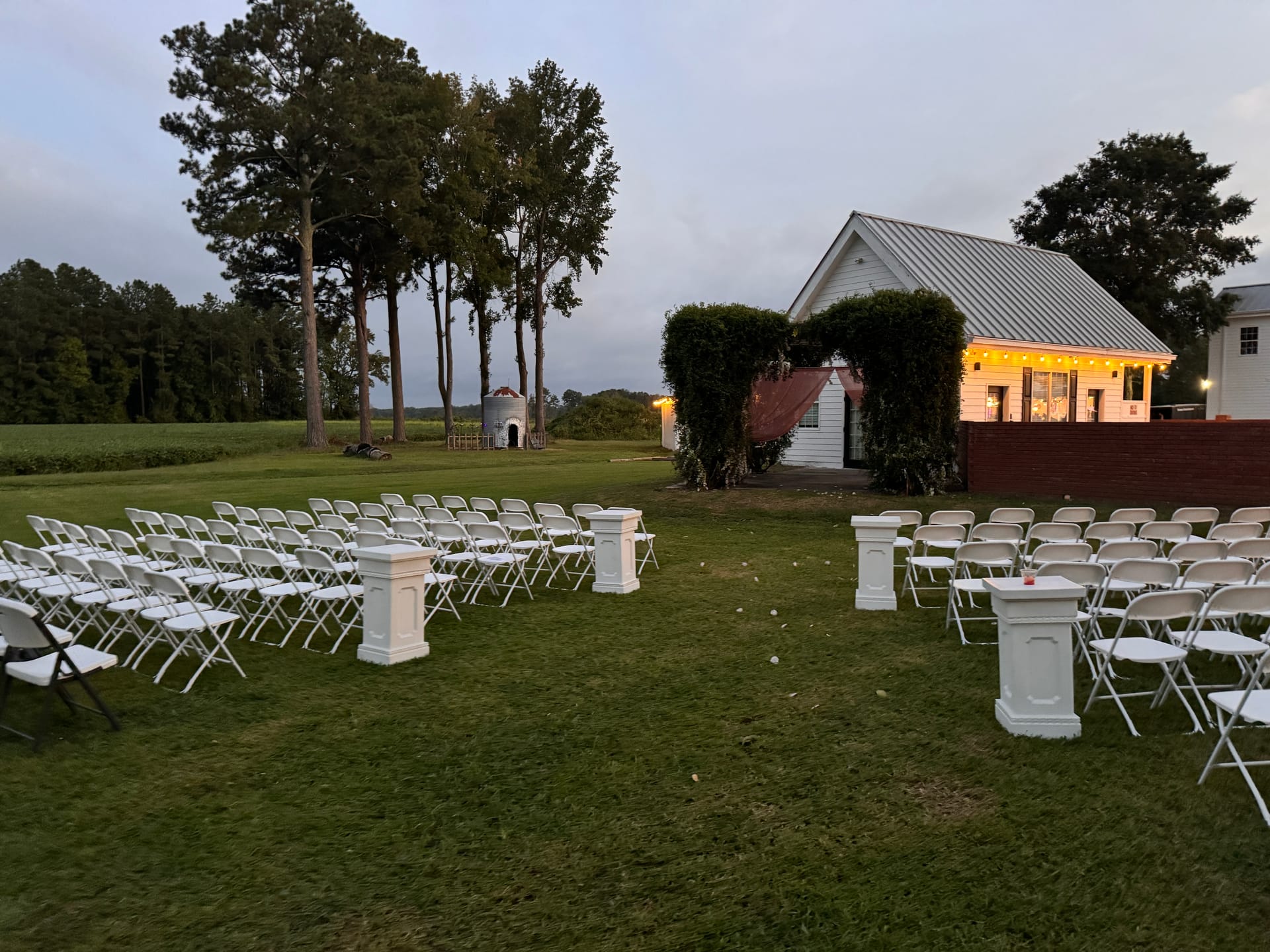 Outdoor wedding ceremony setup at dusk featuring rows of white folding chairs flanking a central aisle with decorative white pillars and a warmly lit historic white farmhouse venue in the background surrounded by tall trees and lush greenery.