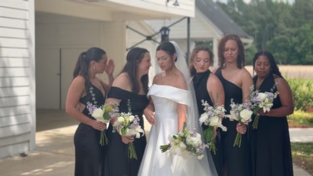 Bride in white gown and veil surrounded by five bridesmaids in black dresses holding purple and white bouquets outside Scarborough House historic estate.