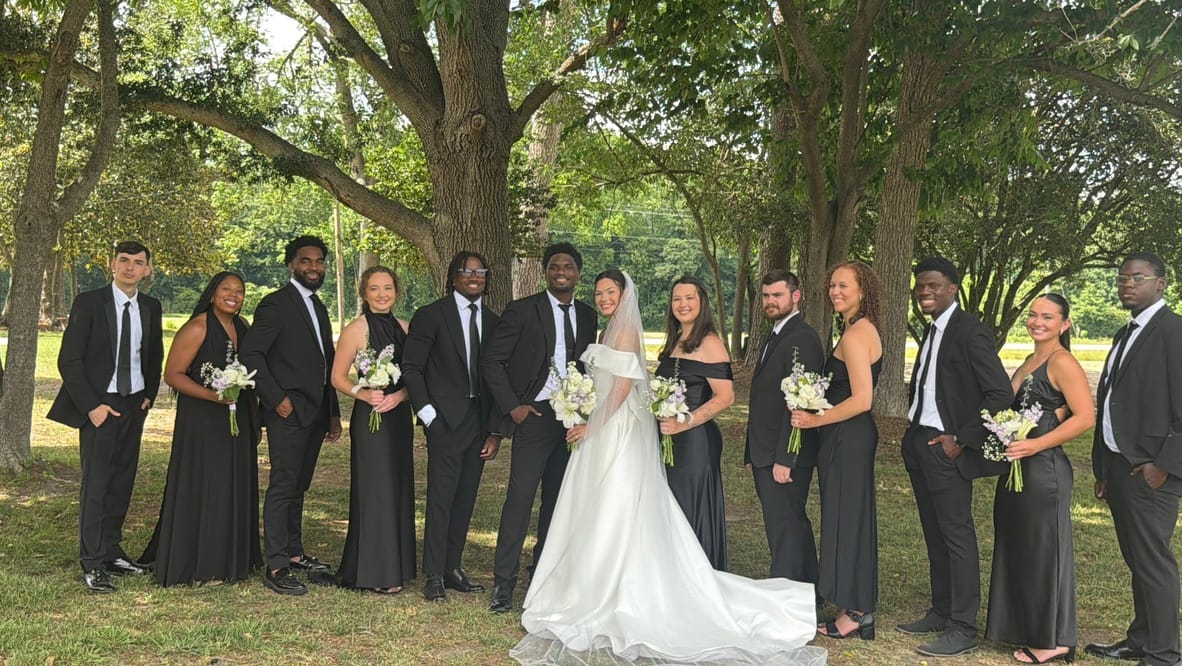 Bride in a white off-shoulder gown and groom in a black suit stand at center surrounded by their wedding party dressed in all black beneath the sweeping oak trees at Scarborough House estate.