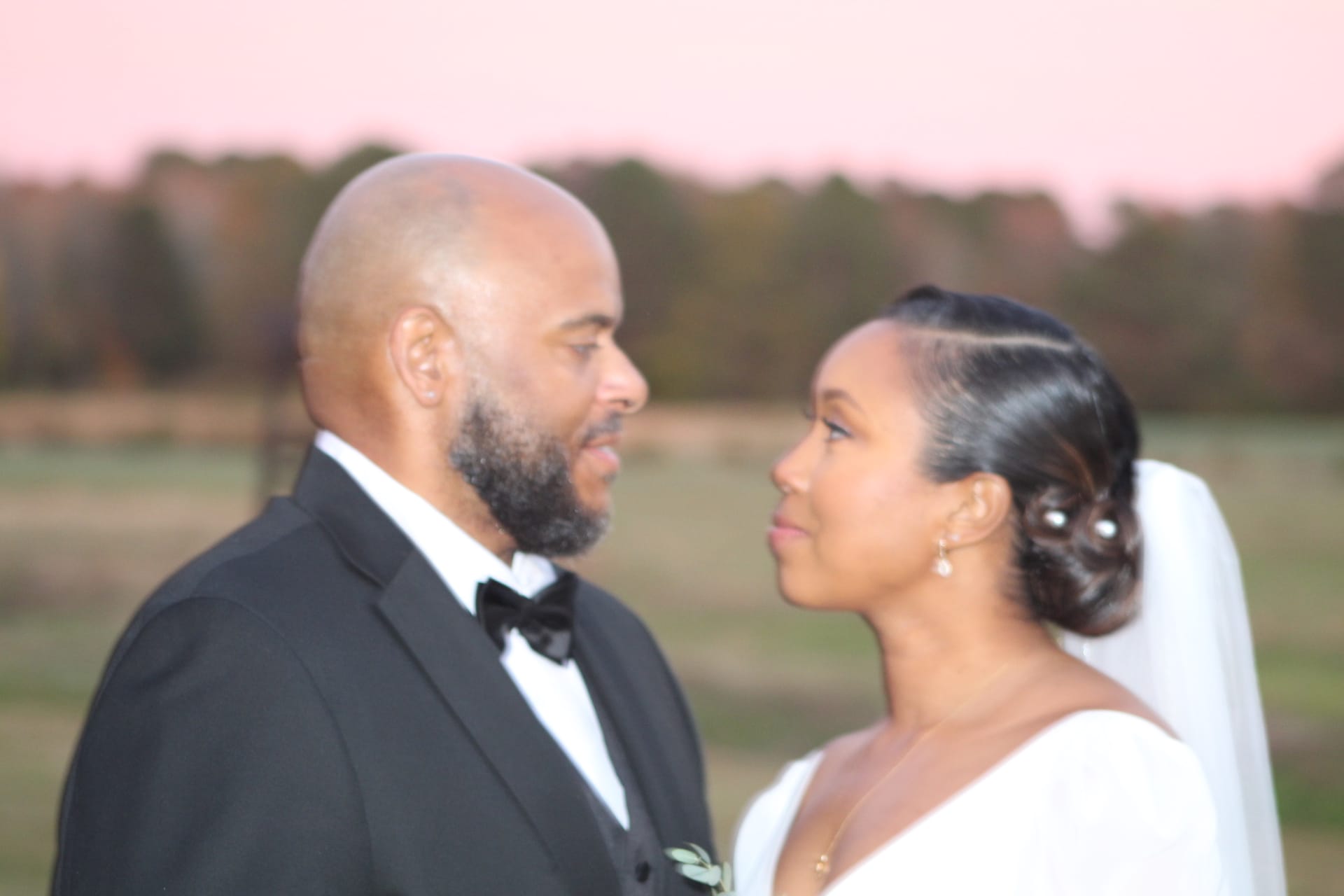 Bride in a white gown and veil gazes at her tuxedo-clad groom beneath a soft pink sunset sky over the open farmland fields at Scarborough House.