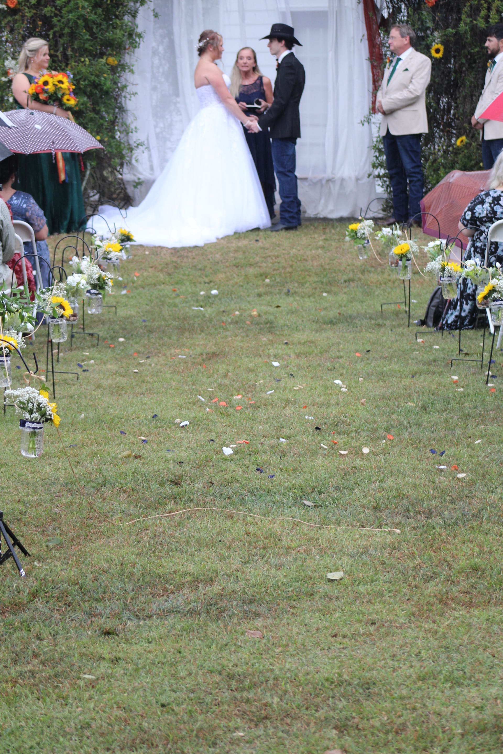 Bride in white ballgown and groom in black cowboy hat exchange vows at an outdoor ceremony on a grass aisle lined with sunflower mason jar arrangements at Scarborough House.