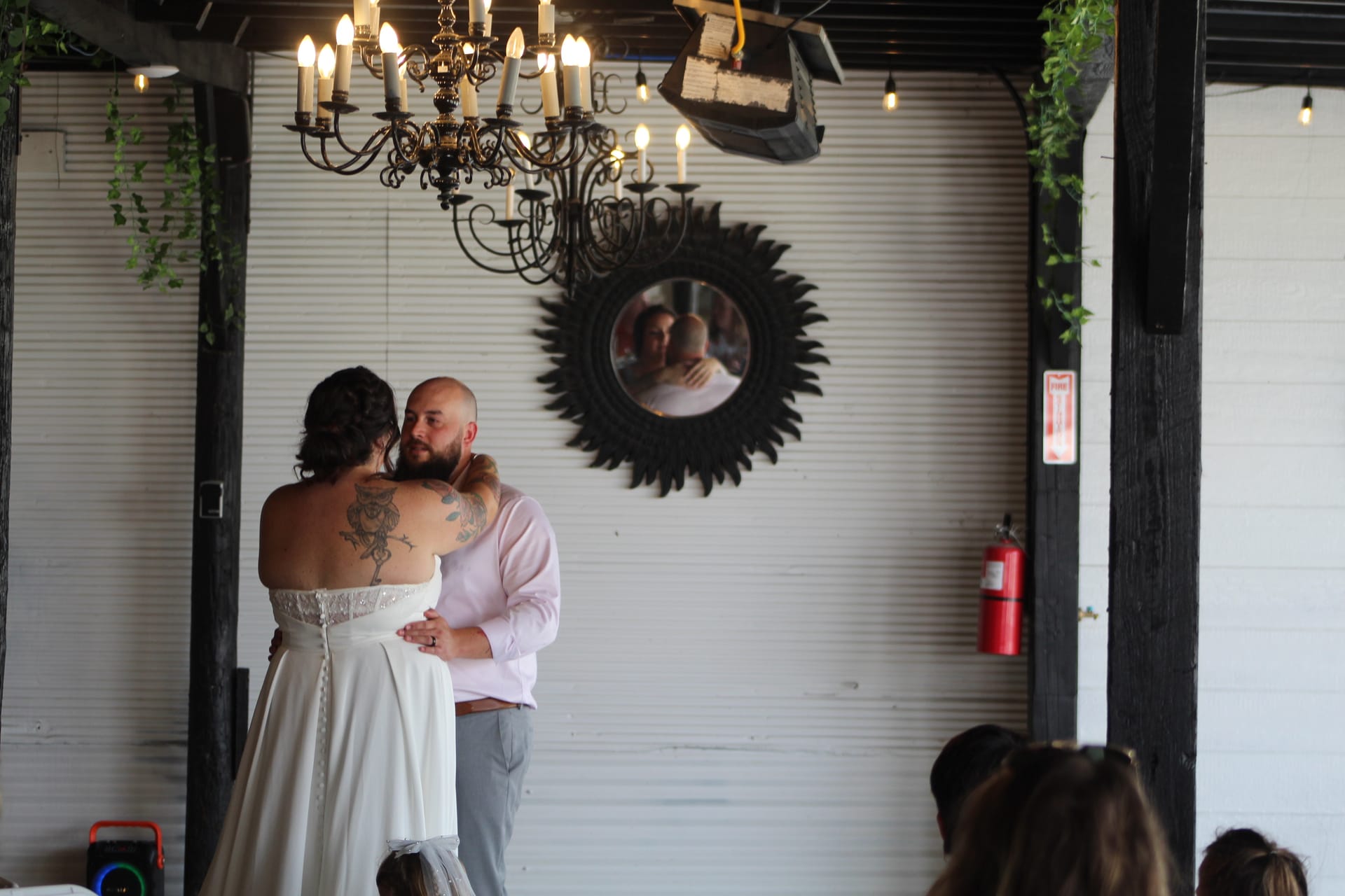 Bride and groom share their first dance beneath a wrought-iron chandelier at Scarborough House, their embrace reflected in a sunburst mirror on the white shiplap wall behind them.