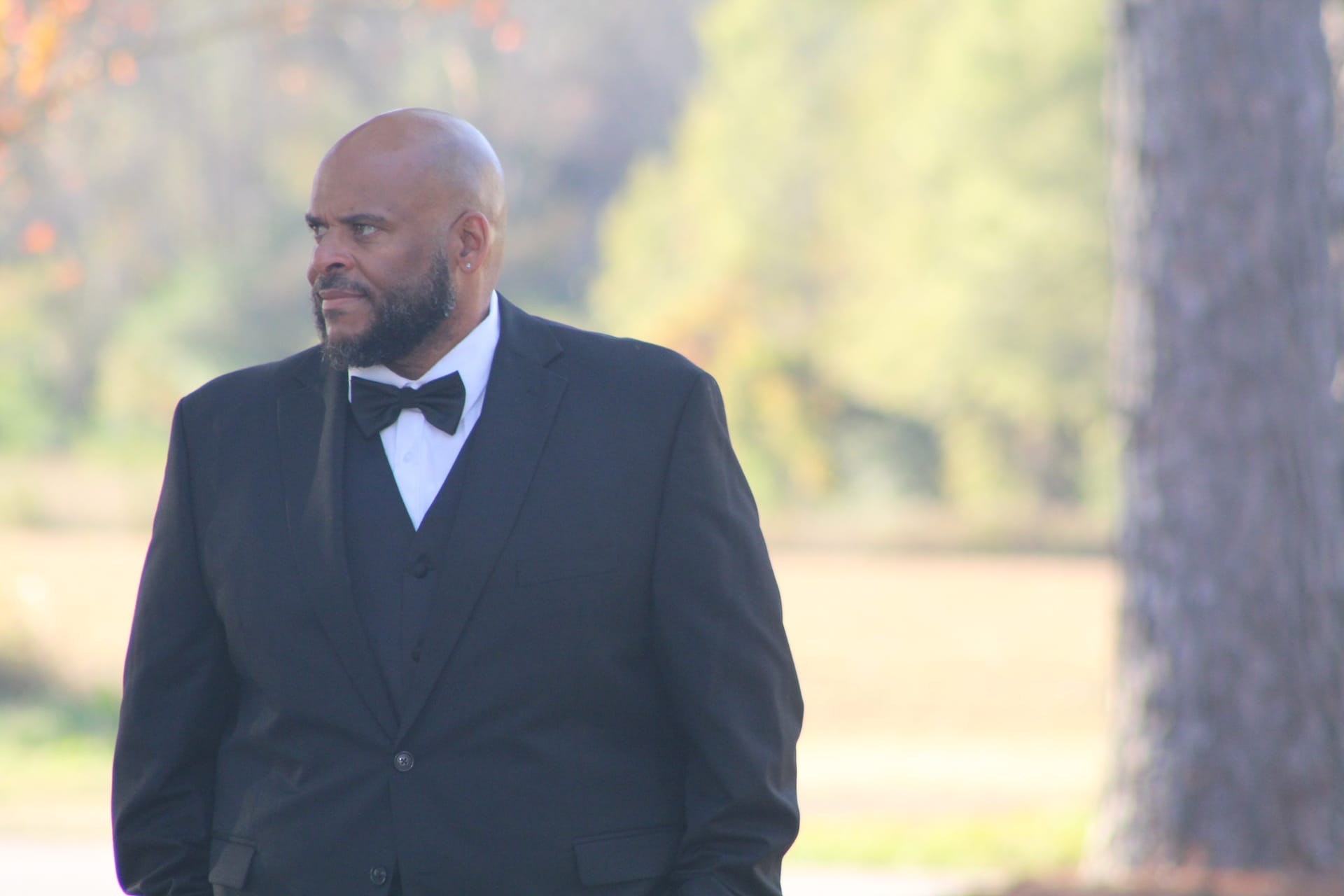 A well-dressed groom in a black tuxedo and bow tie stands outdoors at Scarborough House estate with softly blurred autumn trees and farmland fields in the background.