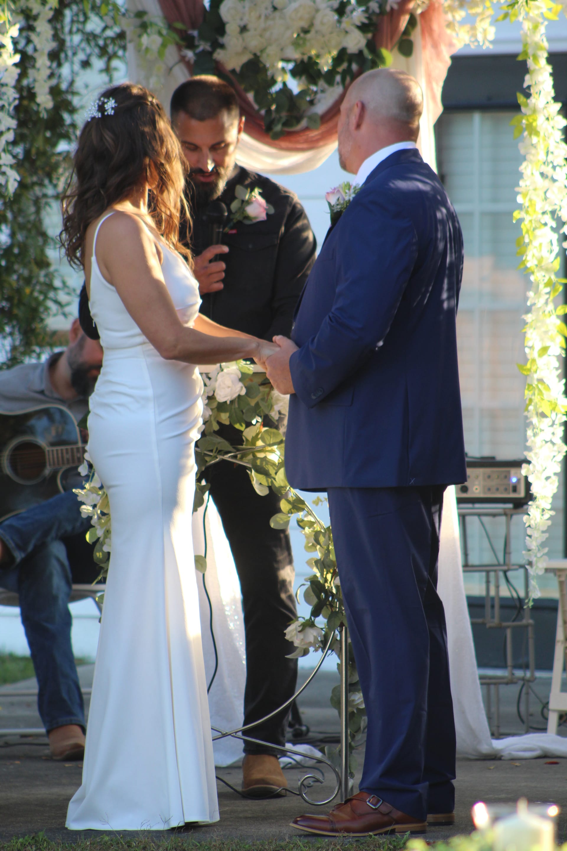 Bride in a fitted white gown and groom in a navy suit hold hands during their outdoor wedding ceremony beneath a floral arch draped with greenery and pink roses at Scarborough House.