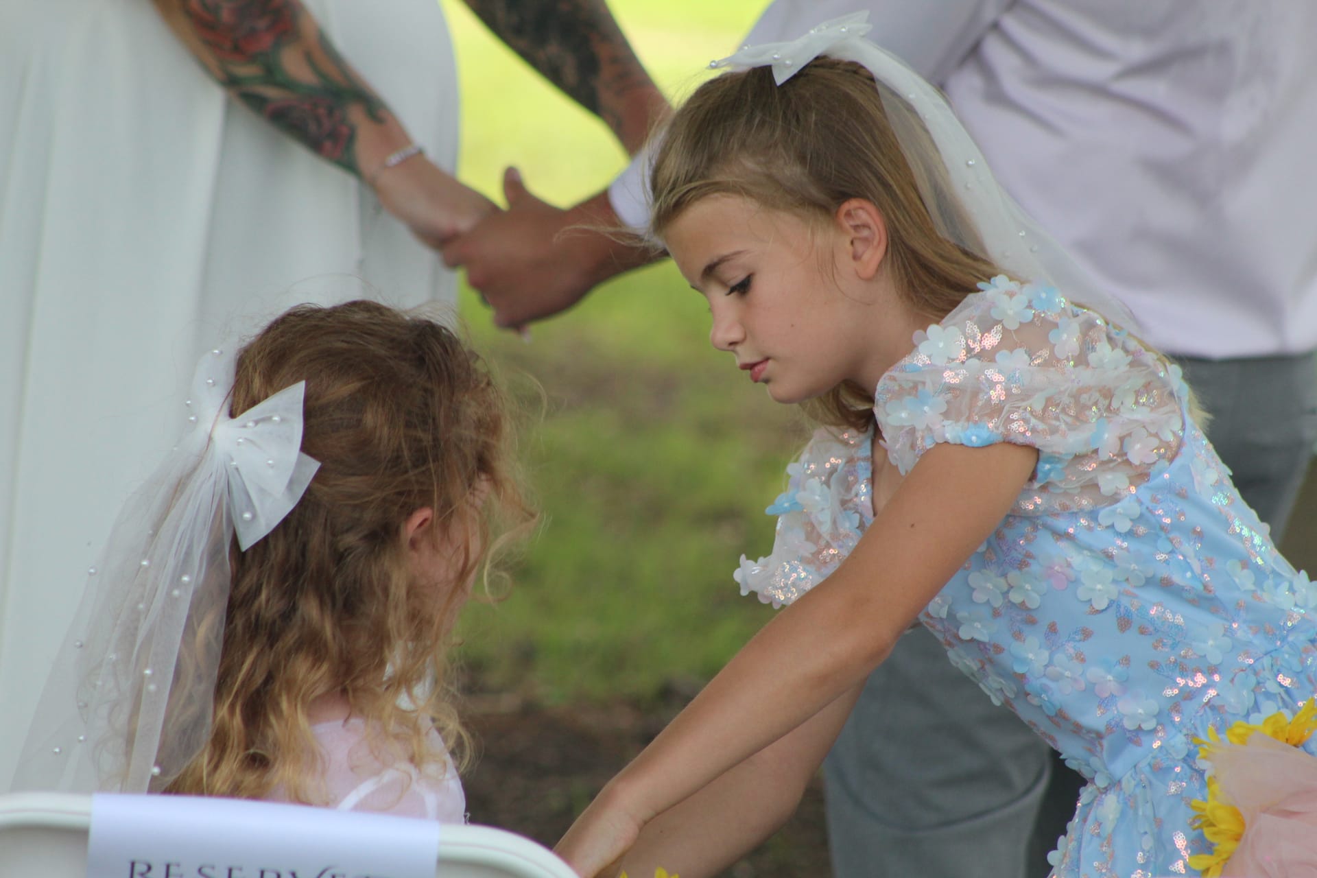 Two young flower girls in white bow veils and floral dresses share a sweet candid moment together during an outdoor wedding ceremony at Scarborough House.