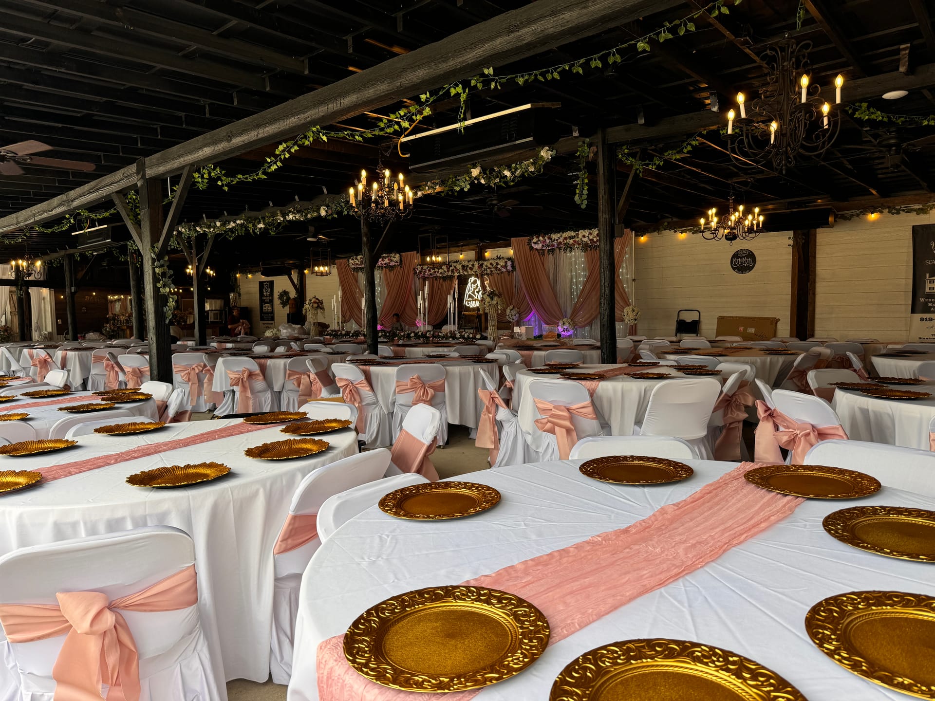 Elegantly decorated grand hall reception space with white linen tables, gold charger plates, blush satin chair sashes and table runners, rustic wood beam ceiling draped with greenery garlands and candle chandeliers, and a floral blush drapery backdrop stage at Scarborough House.
