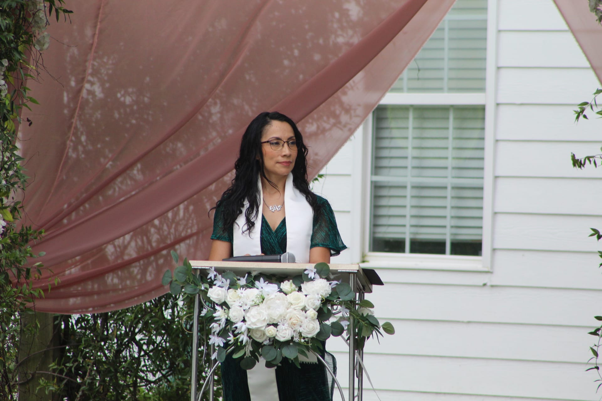 A wedding officiant in a green dress and white stole speaks at a floral-decorated podium beneath dusty rose draped fabric at an outdoor ceremony.