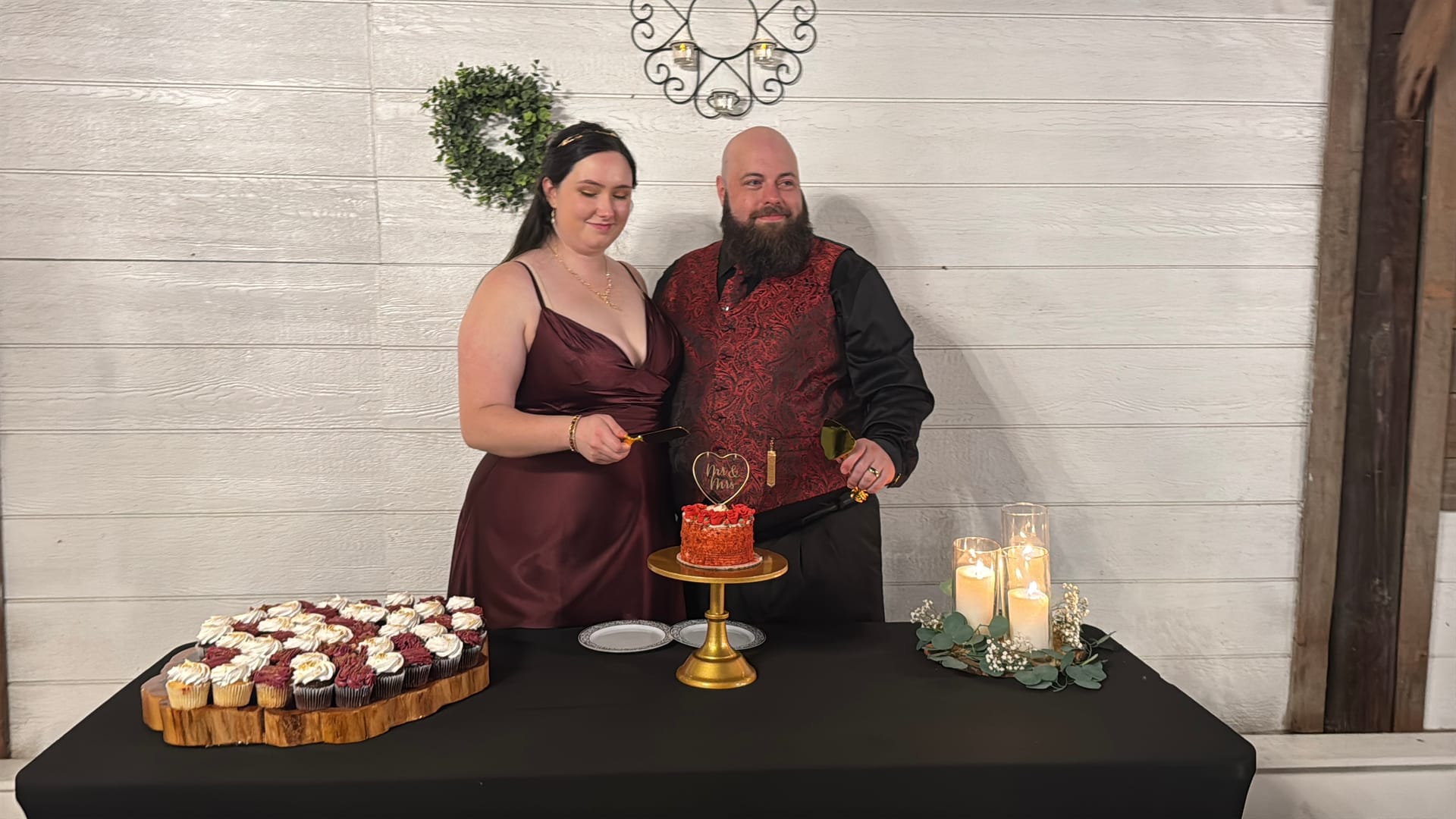 Bride and groom in coordinating burgundy attire prepare to cut their red wedding cake on a gold cake stand surrounded by cupcakes and candlelight at Scarborough House.