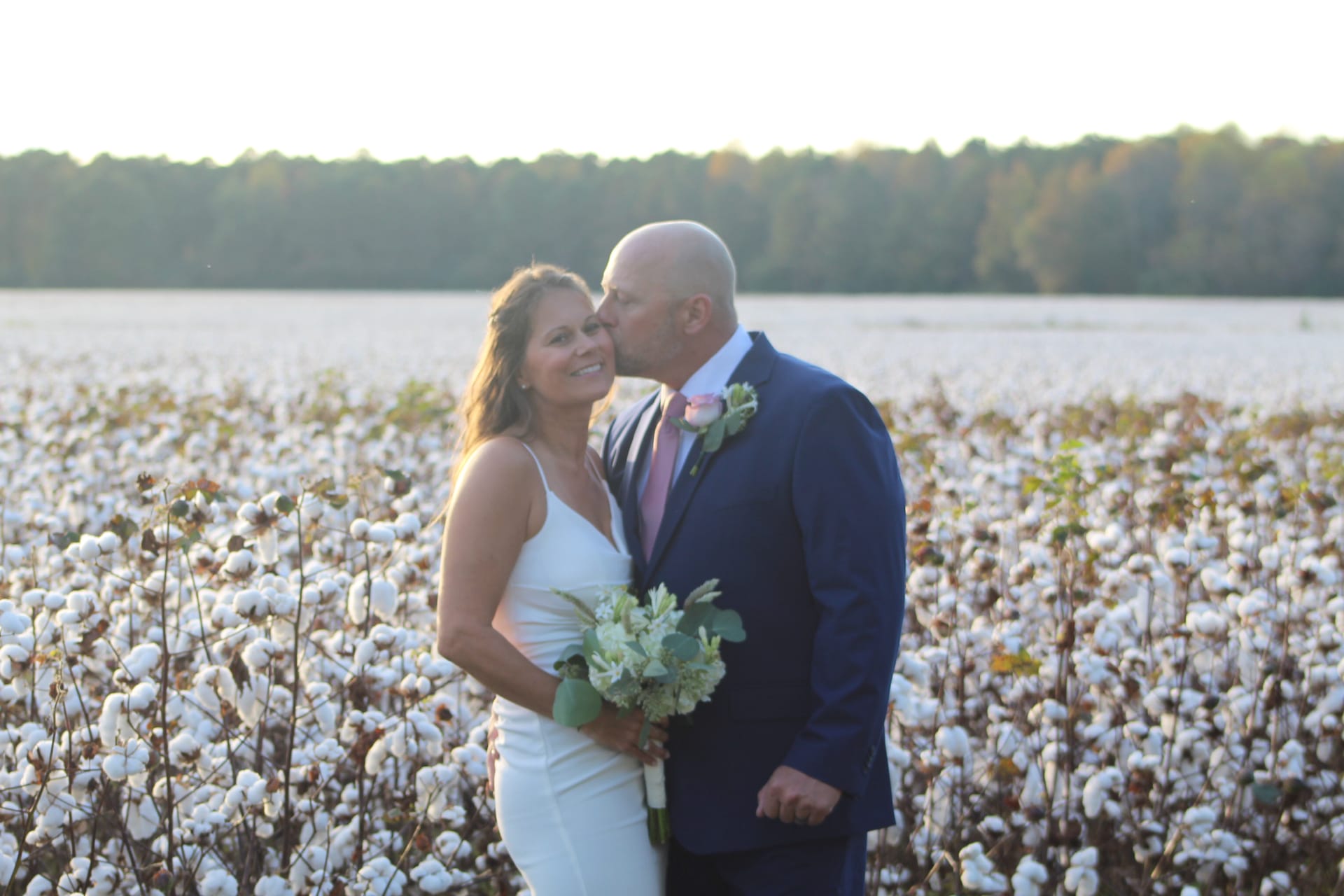 Bride in white dress holding a bouquet smiles as groom in navy suit kisses her cheek in a sunlit cotton field at Scarborough House.