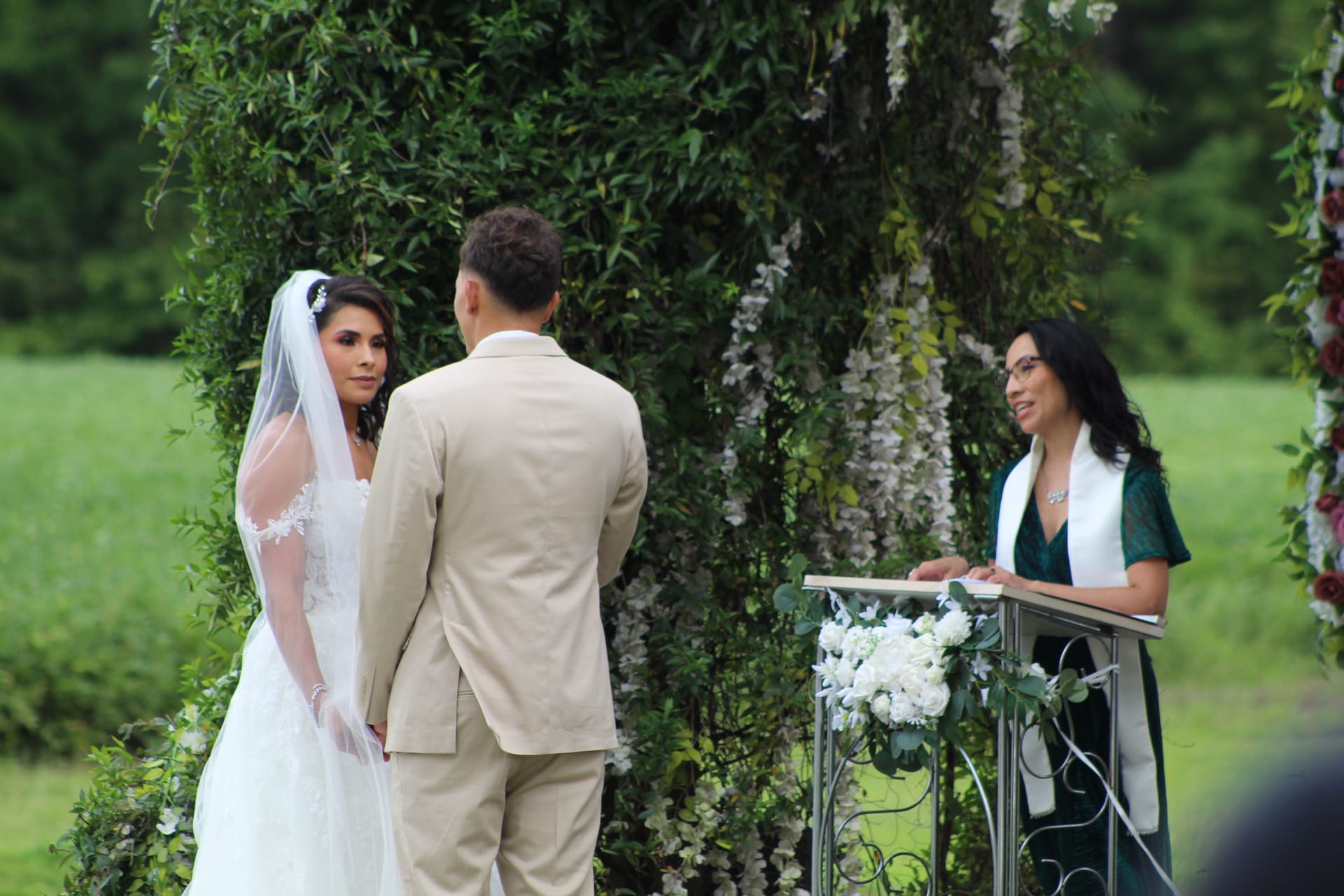 Bride in white lace gown and veil gazes at groom during outdoor wedding ceremony beneath a lush floral arch while officiant speaks at a podium adorned with white flowers.