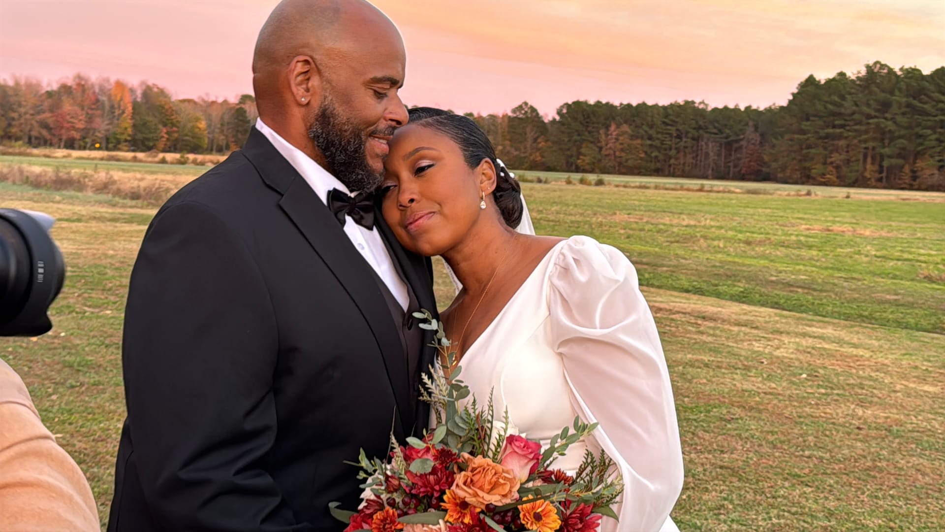Bride resting her head on groom's shoulder in a sun-drenched autumn field at Scarborough House, holding a warm-toned floral bouquet beneath a dramatic pink and orange sunset sky.