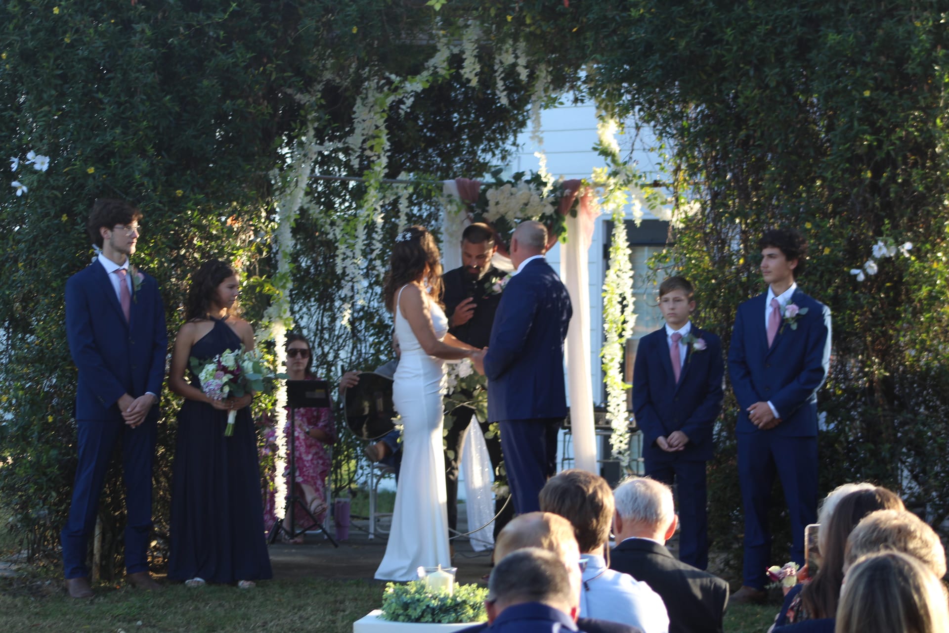 Bride and groom exchange vows under a white floral arch surrounded by lush greenery as their wedding party looks on during an outdoor ceremony at Scarborough House.