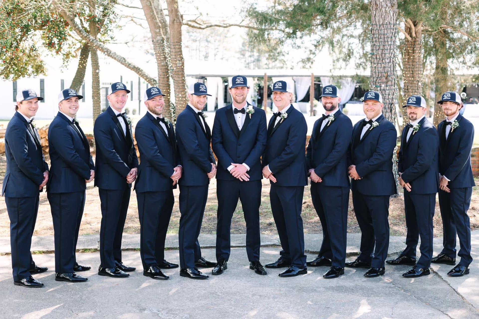 Groom and ten groomsmen in matching navy tuxedos and custom caps posed in a symmetrical line outdoors among tall trees at Scarborough House wedding venue.