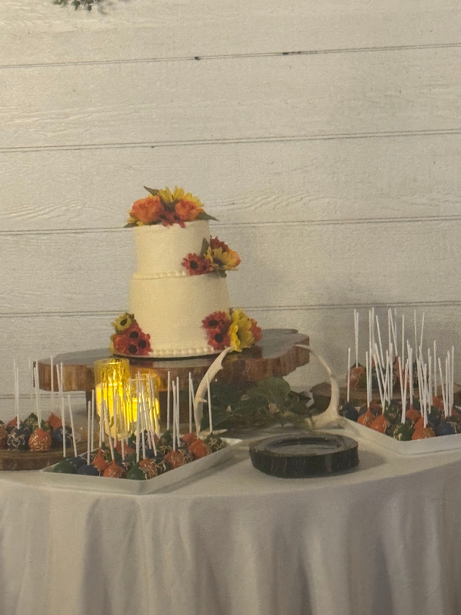 A two-tier white wedding cake decorated with autumn sunflowers and orange roses displayed on a rustic wood slice stand alongside cake pops and chocolate-dipped strawberries at Scarborough House.