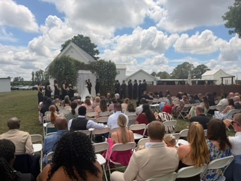 Guests seated in white chairs on the ceremony lawn watch a wedding party gathered under a white arch adorned with greenery at Scarborough House historic estate.