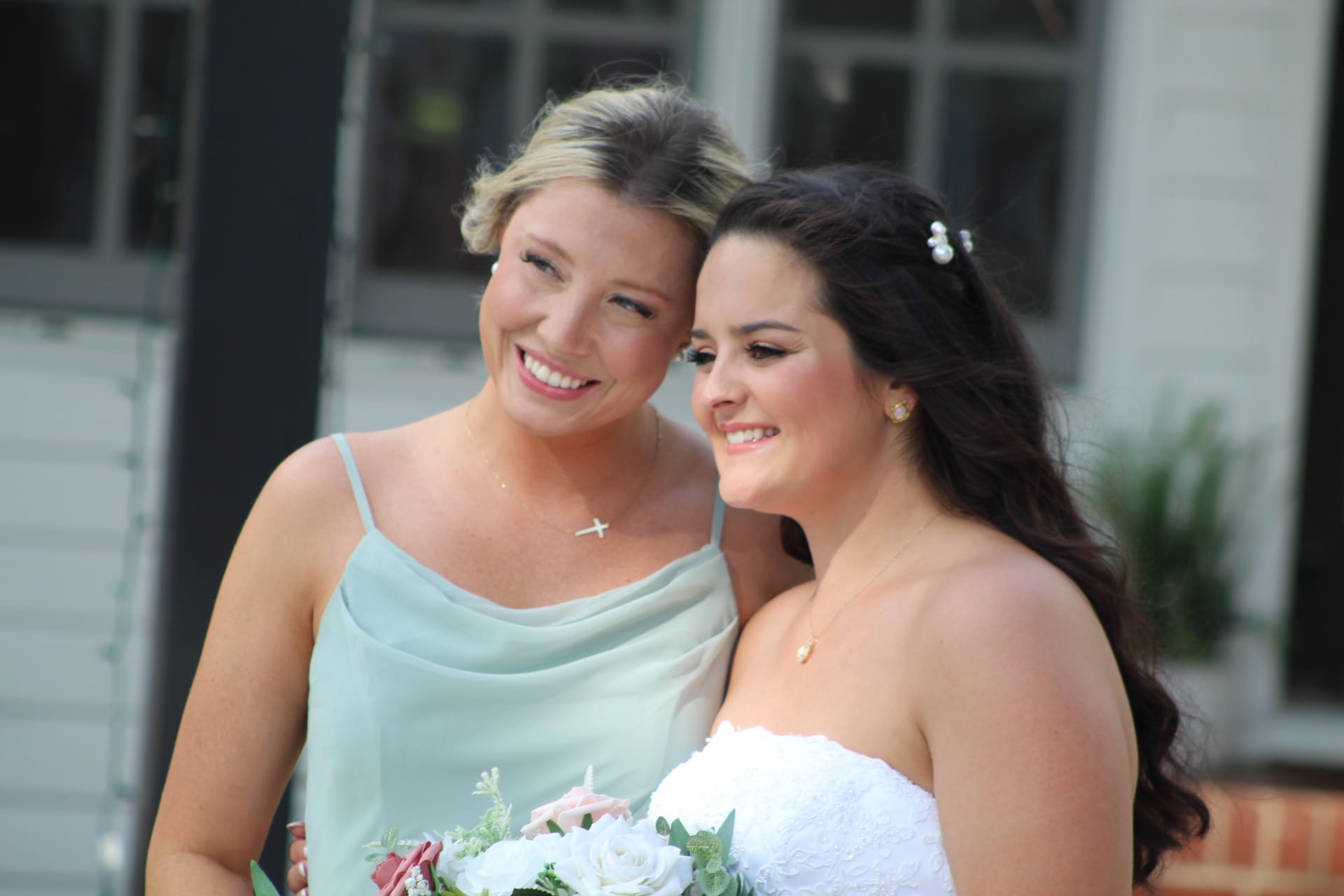 Smiling bride in a white strapless gown and her bridesmaid in a sage green dress lean together holding a floral bouquet outside a historic white manor house.