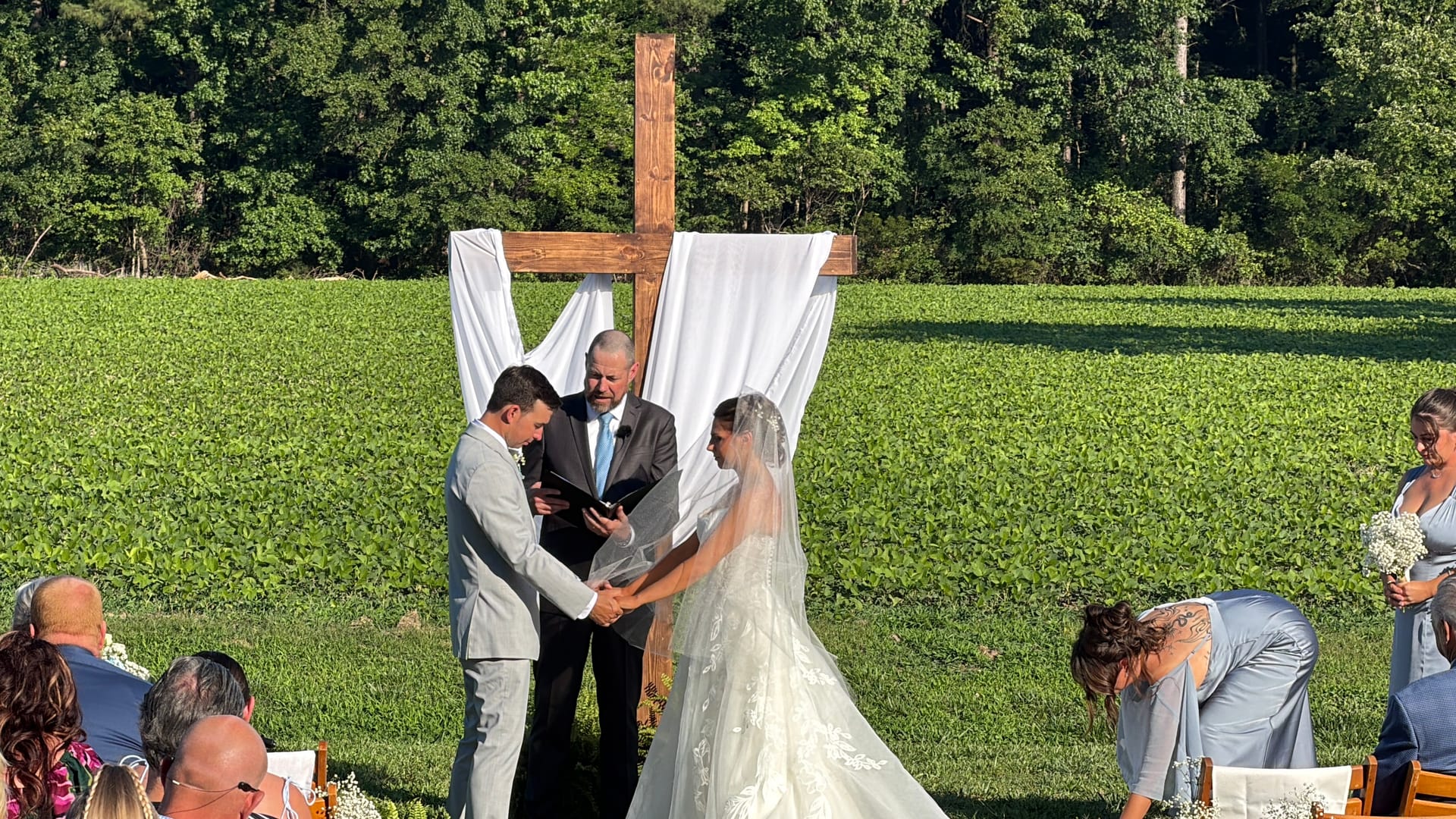 Bride and groom hold hands during their outdoor wedding ceremony beneath a draped wooden cross with a lush green field and wooded treeline in the background at Scarborough House estate.