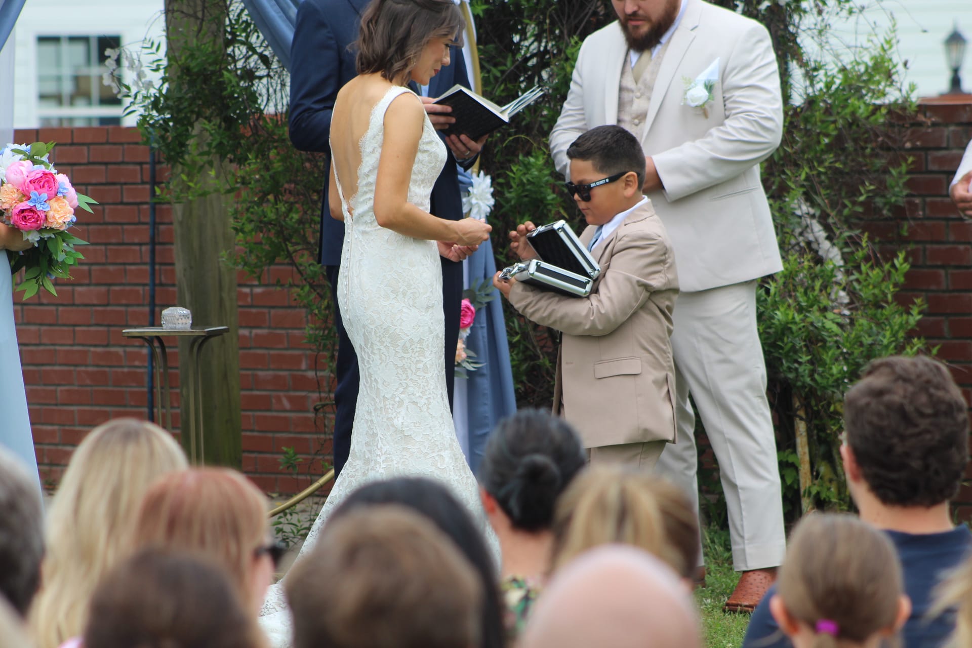 A young ring bearer in a tan suit and sunglasses presents the ring case during an outdoor wedding ceremony at Scarborough House as the bride in a backless lace gown looks on.