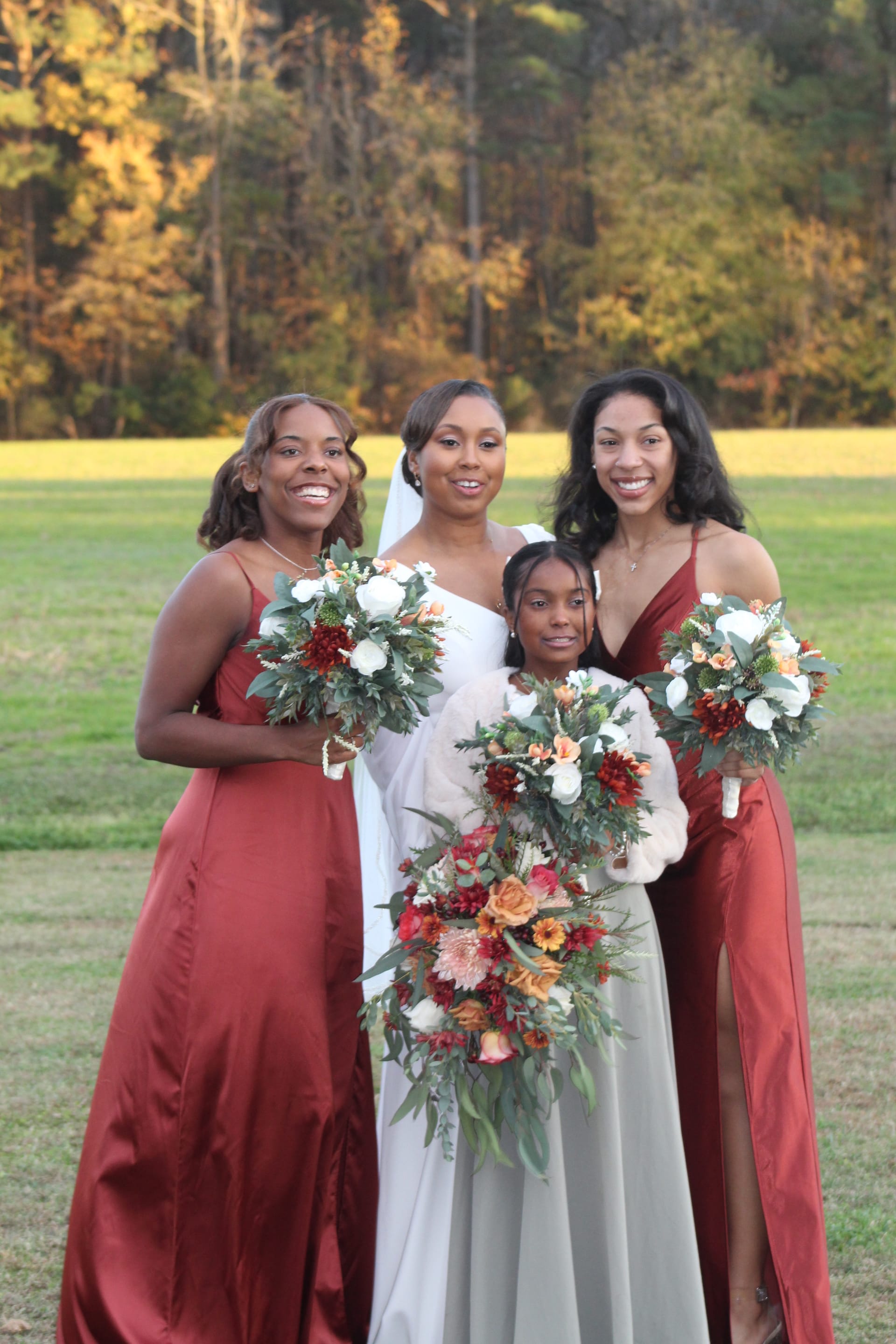 Bride in white gown and veil poses with two bridesmaids in rust-red dresses and a young attendant holding lush autumn bouquets on the open farmland lawn at Scarborough House.