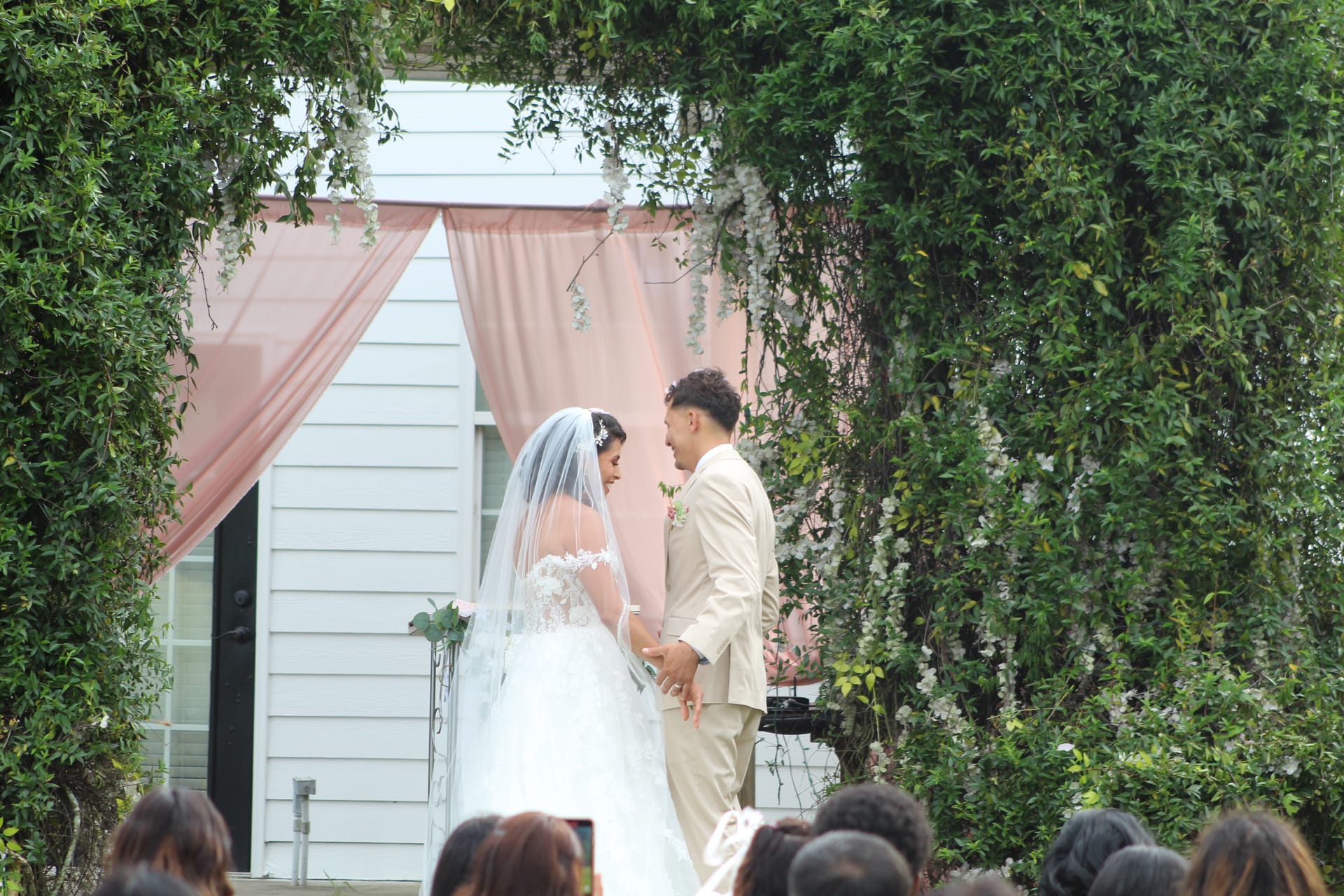 Bride in lace gown and veil holds hands with groom in tan suit at an outdoor ceremony altar framed by a verdant green arch and soft blush curtains at Scarborough House.