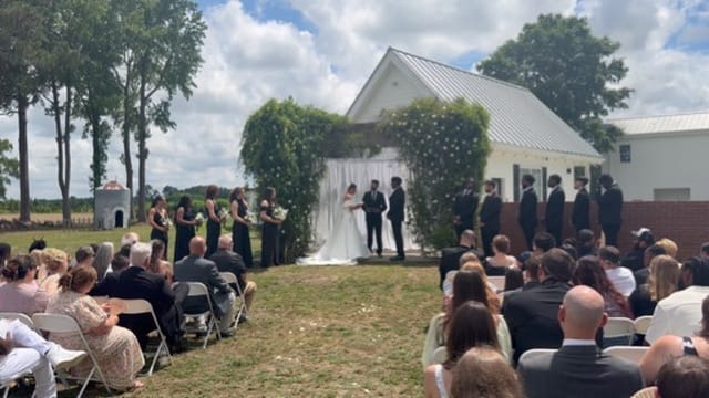 Outdoor wedding ceremony on the lawn at Scarborough House with bride and groom at an ivy-covered arch flanked by bridesmaids in black and groomsmen in suits while guests look on.