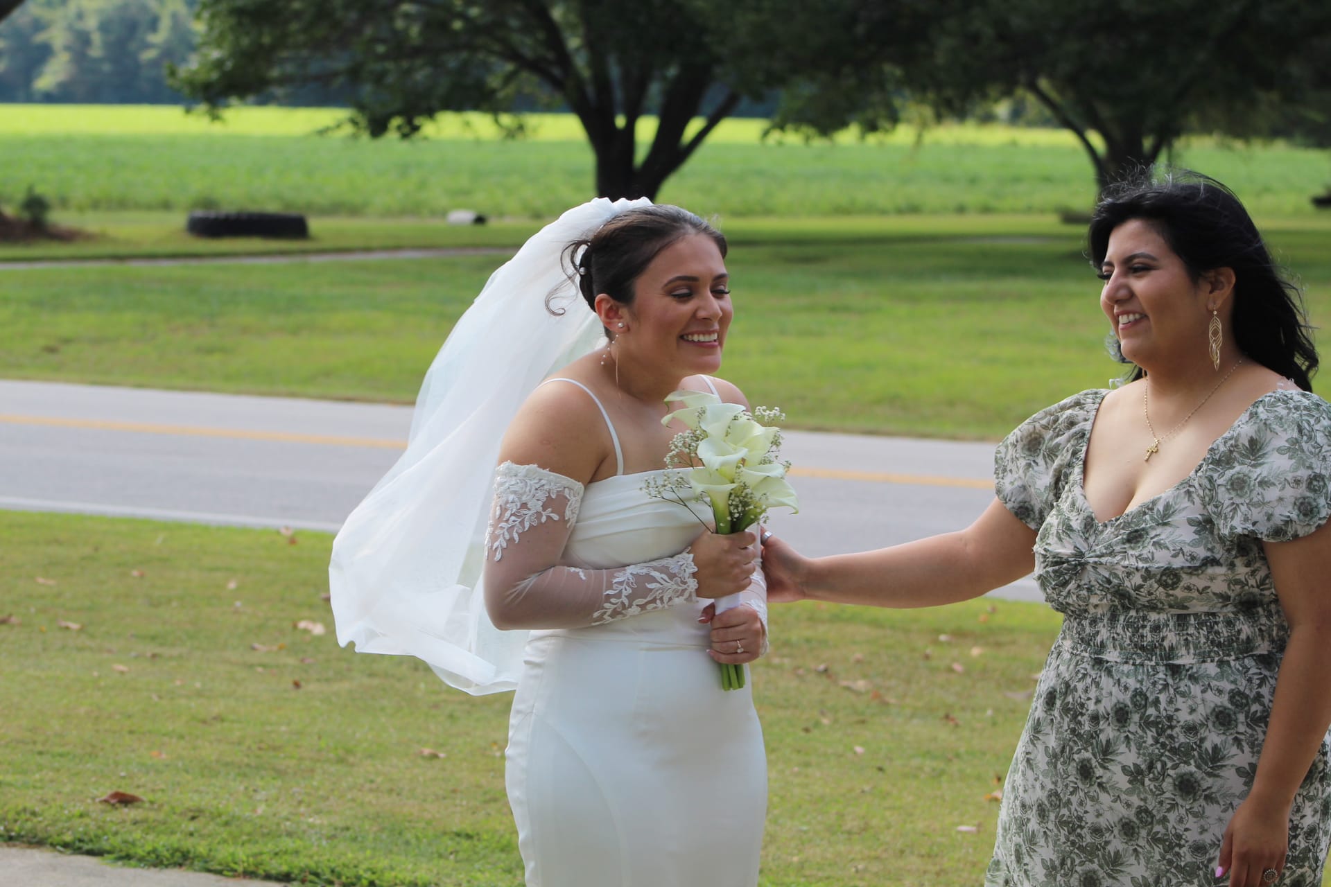 Smiling bride in a white gown and veil holding a white calla lily bouquet shares a joyful moment with a guest in a floral dress against a lush green farmland backdrop.
