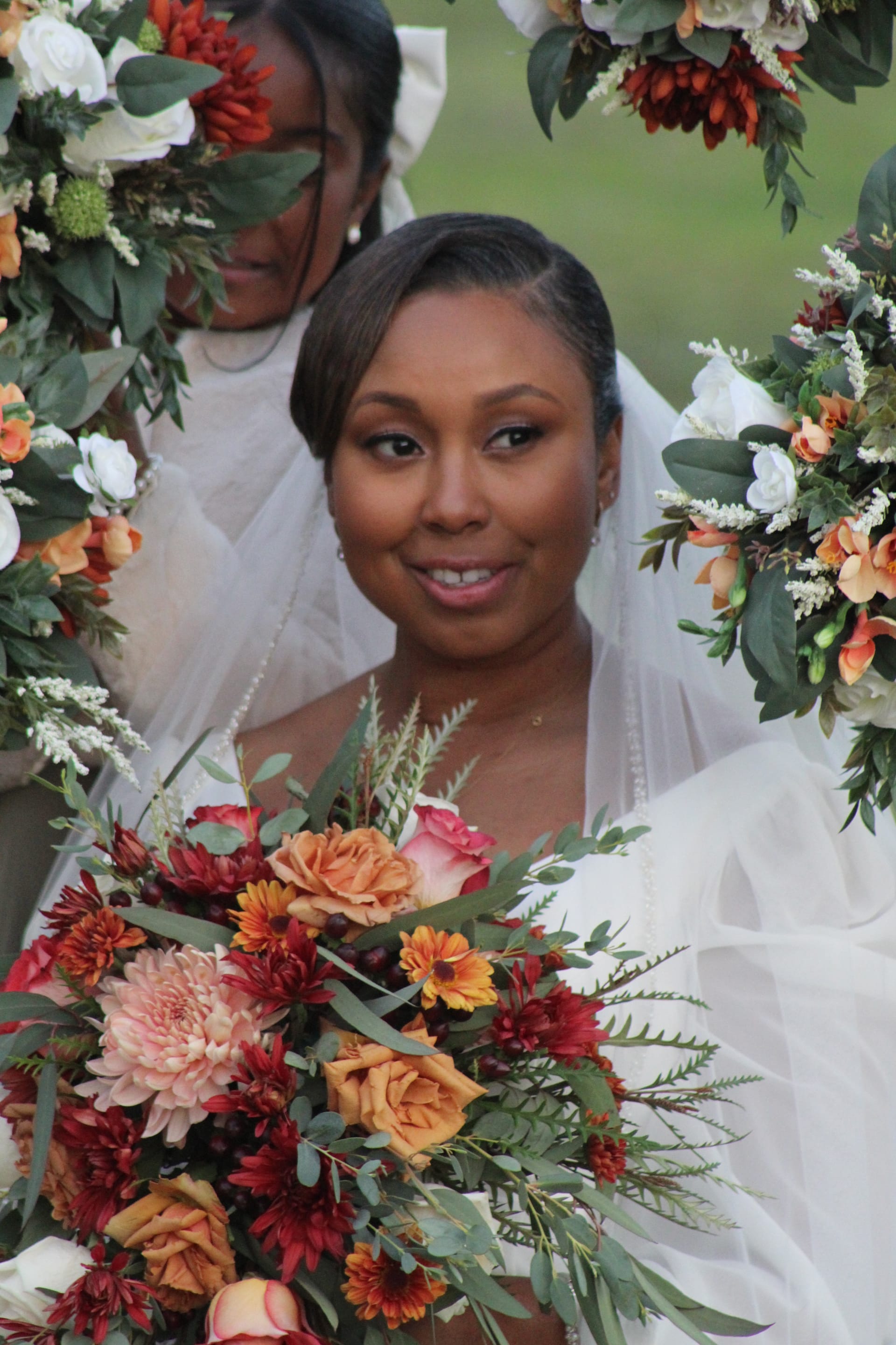 Bride in a white gown and veil smiles warmly while surrounded by rich autumn-toned floral arrangements of burgundy, orange, and blush blooms at an outdoor garden ceremony.