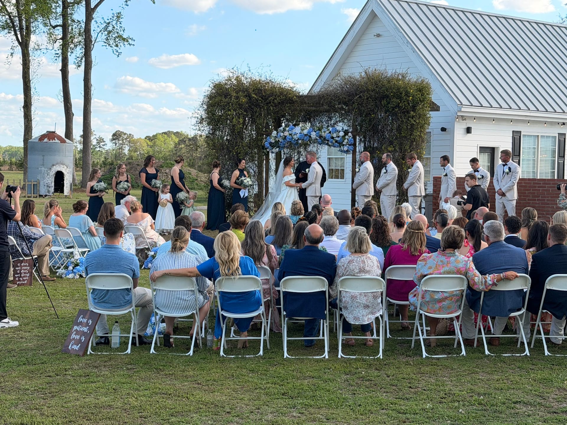 Guests seated in white folding chairs watch a bride and groom exchange vows beneath a lush greenery and blue floral arch on the ceremony lawn at Scarborough House historic estate.