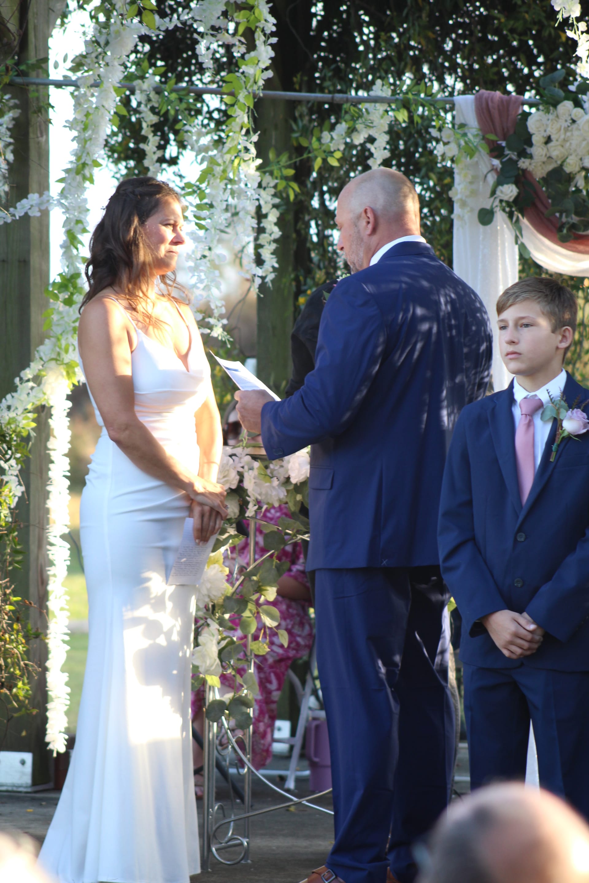 Groom reads handwritten vows to his bride in a white gown beneath a lush floral arch during an outdoor wedding ceremony at Scarborough House.