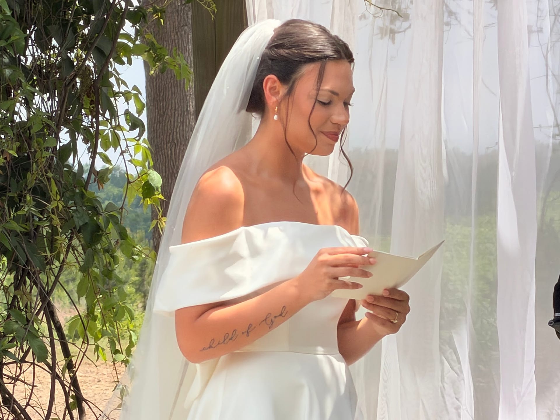 Bride in an off-shoulder white gown and veil reads her wedding vows during an outdoor ceremony surrounded by lush greenery at Scarborough House estate.