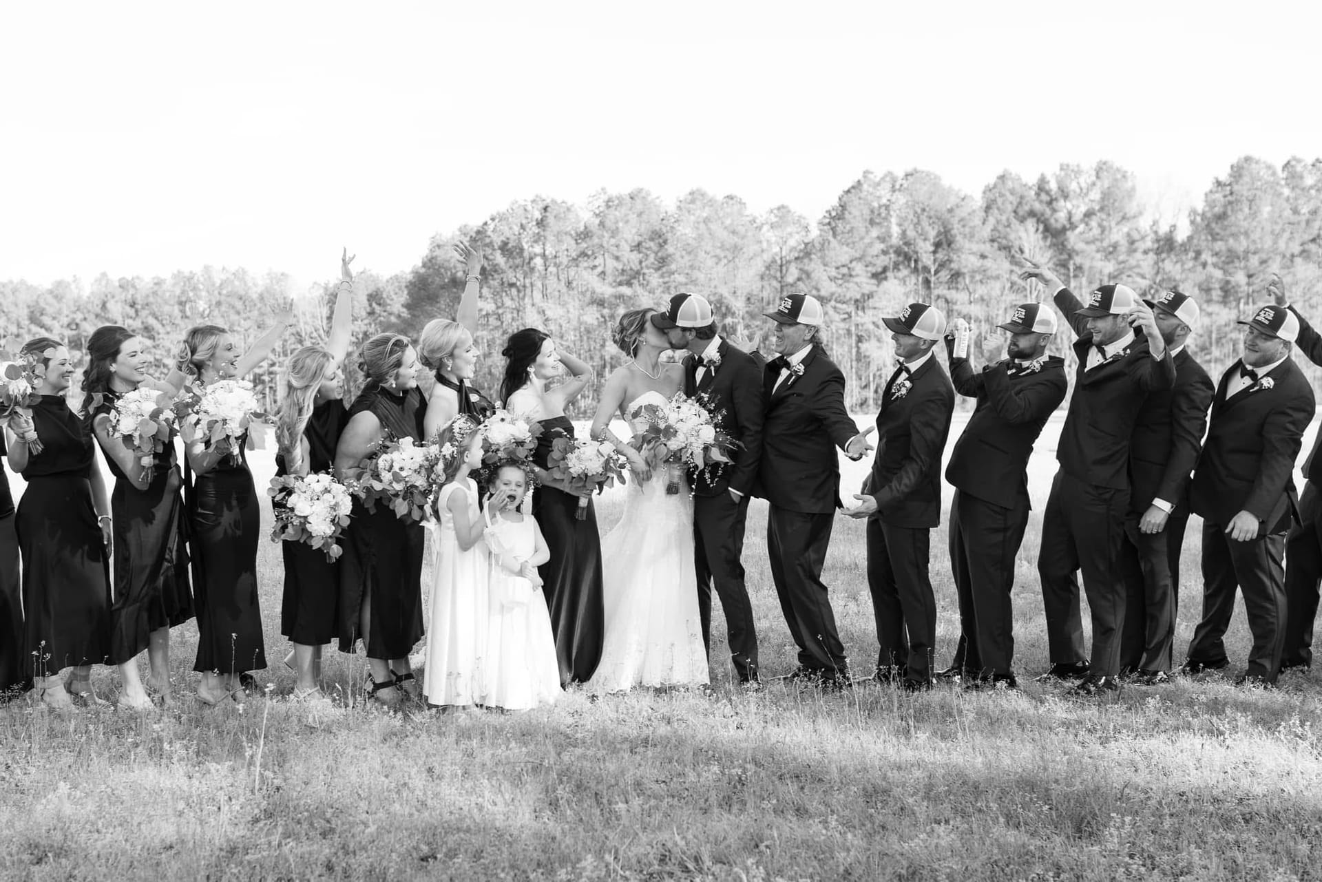 Bride and groom share a kiss surrounded by their full wedding party in a sun-drenched field at Scarborough House, with bridesmaids holding bouquets and groomsmen tipping their caps while a flower girl looks on with delight.