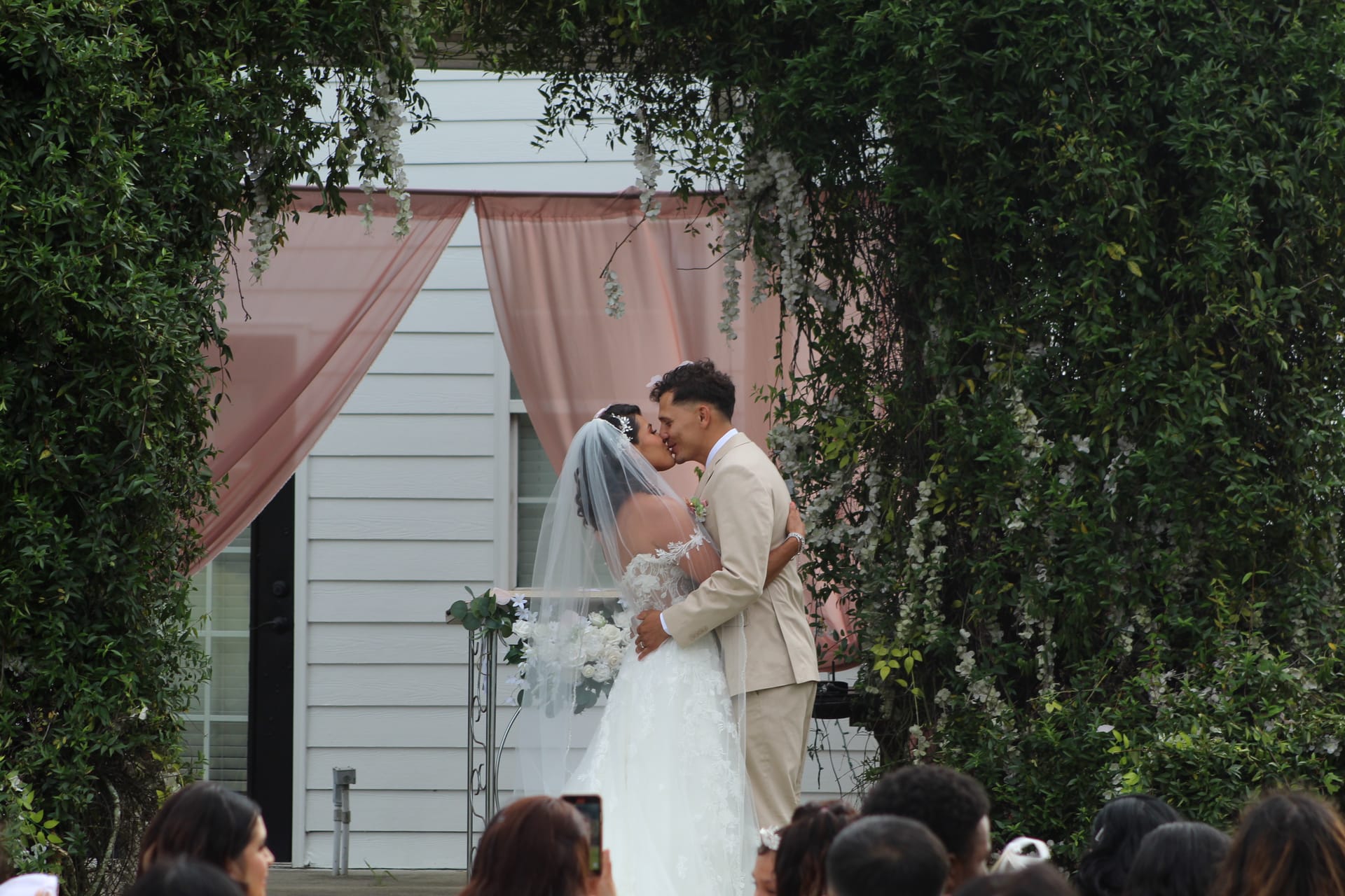Bride and groom share their first kiss beneath a lush ivy-covered arch with blush draping at Scarborough House historic estate wedding venue as guests look on.