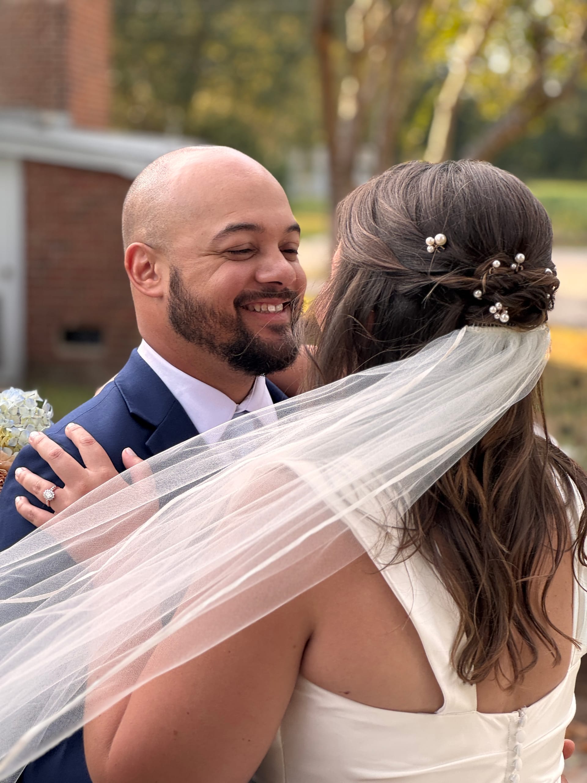 A smiling groom in a navy suit embraces his bride in a white gown and veil adorned with pearl hair pins during their first look outdoors at Scarborough House.
