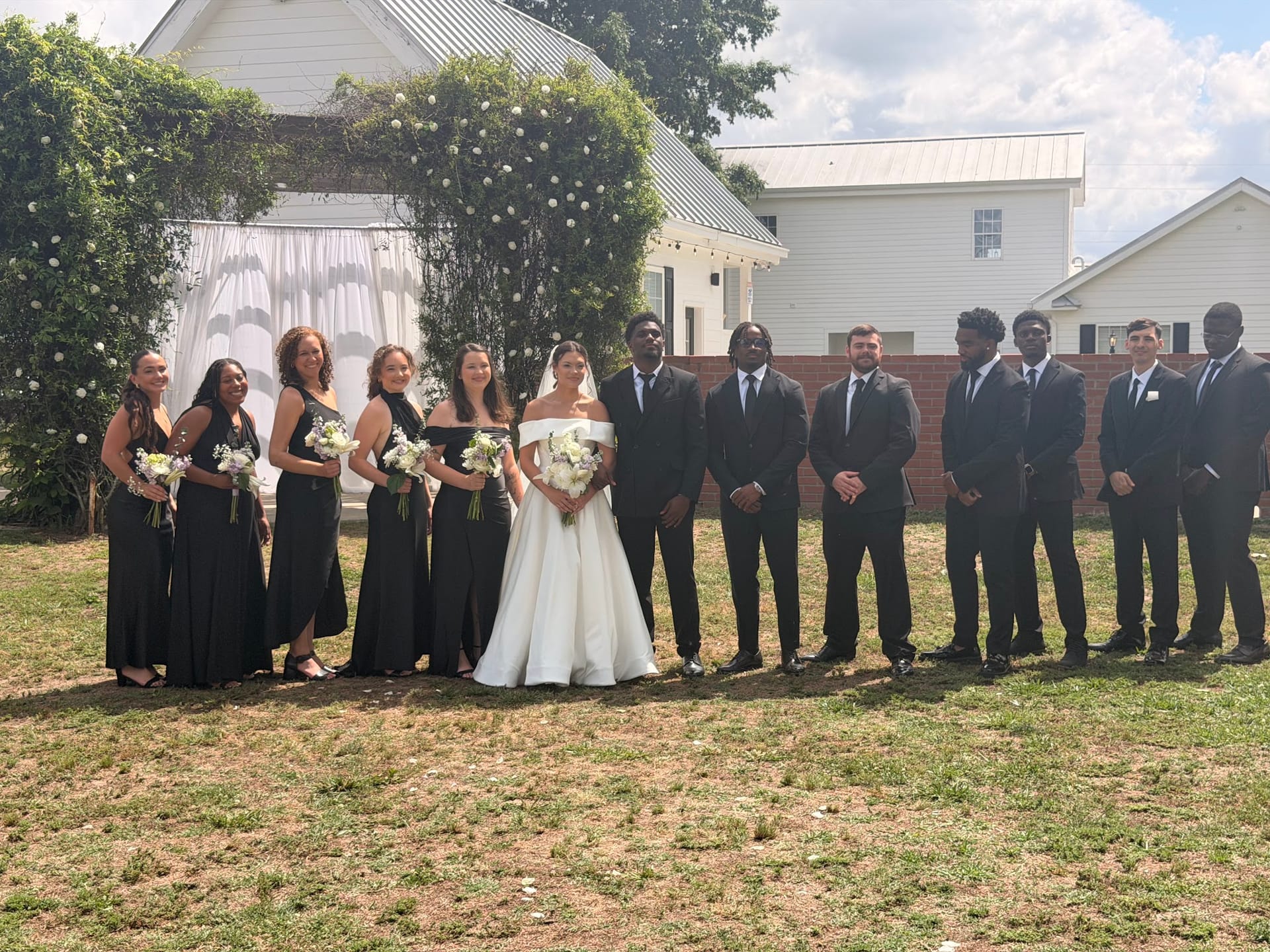 Bride in white ball gown stands with bridesmaids in black dresses and groomsmen in dark suits on the ceremony lawn in front of a floral arch at Scarborough House historic estate.