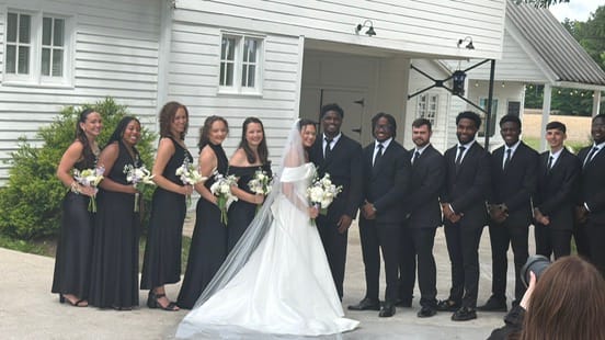 Bride in white gown and veil poses with bridesmaids in black dresses and groomsmen in black suits in front of a white historic farmhouse wedding venue.