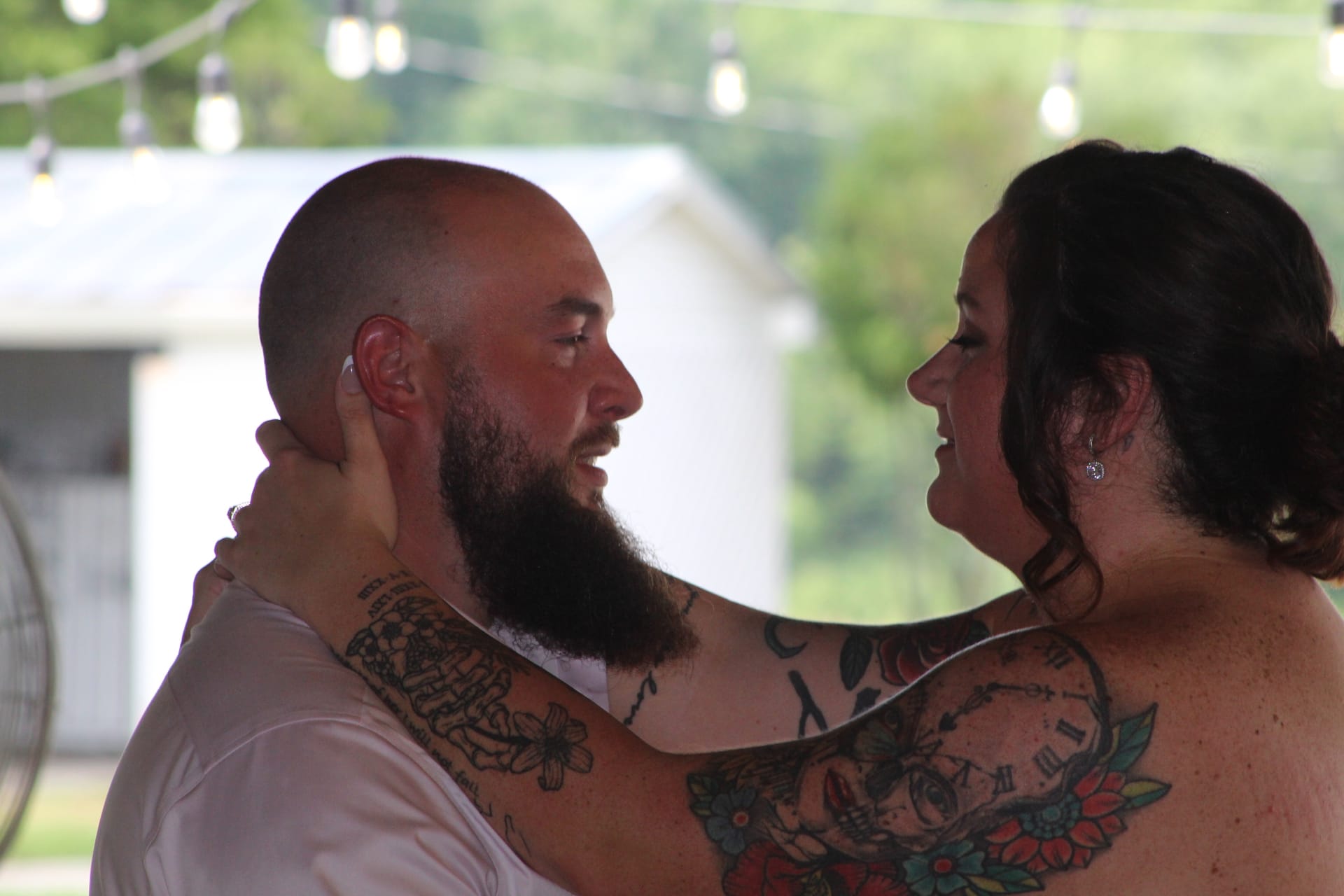 A tattooed bride smiles warmly at her bearded groom during their first dance under string lights at Scarborough House wedding venue.