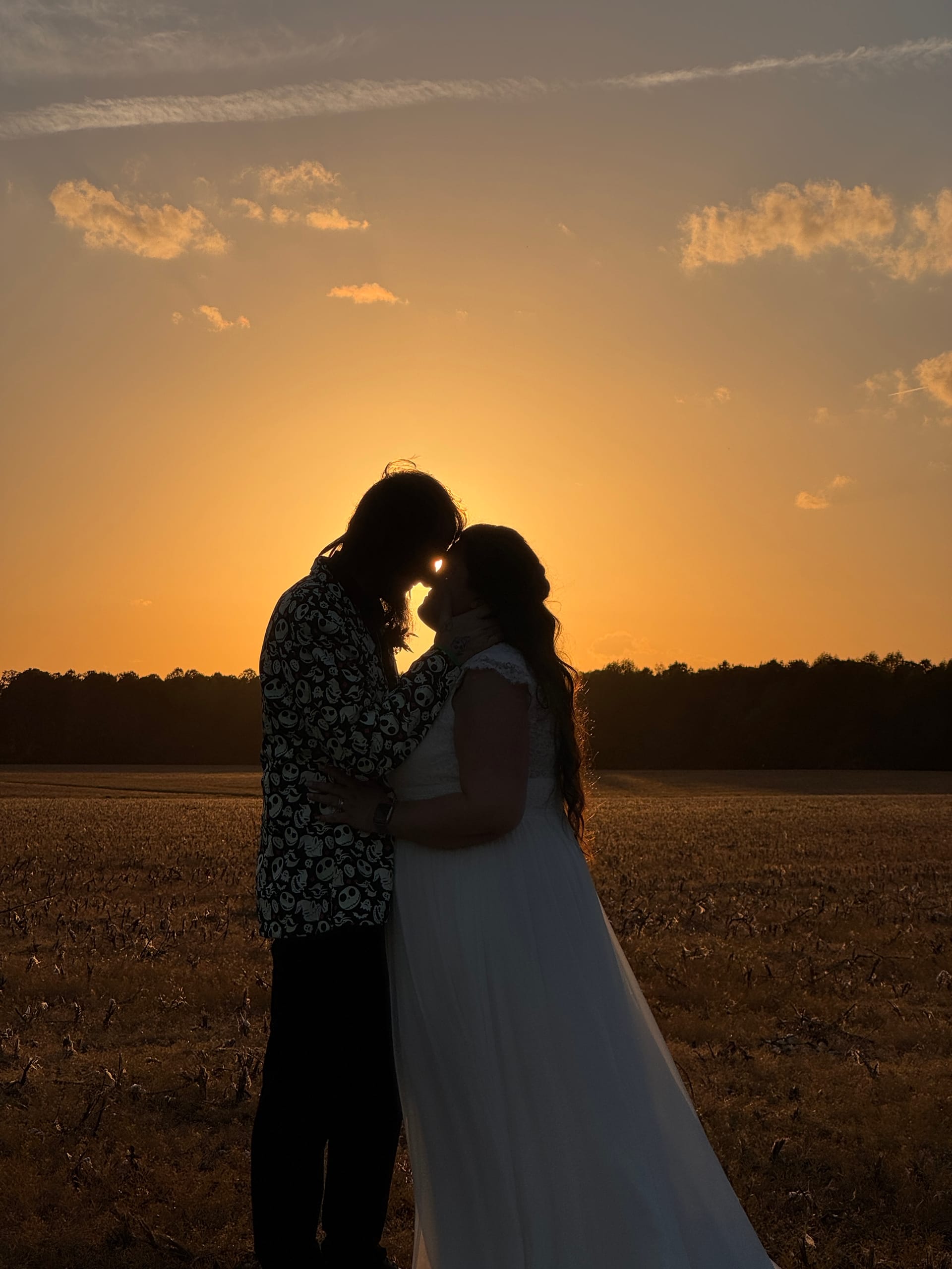 Silhouette of a bride in a white gown and groom in a patterned jacket touching foreheads tenderly against a glowing orange sunset over open farmland fields at Scarborough House.