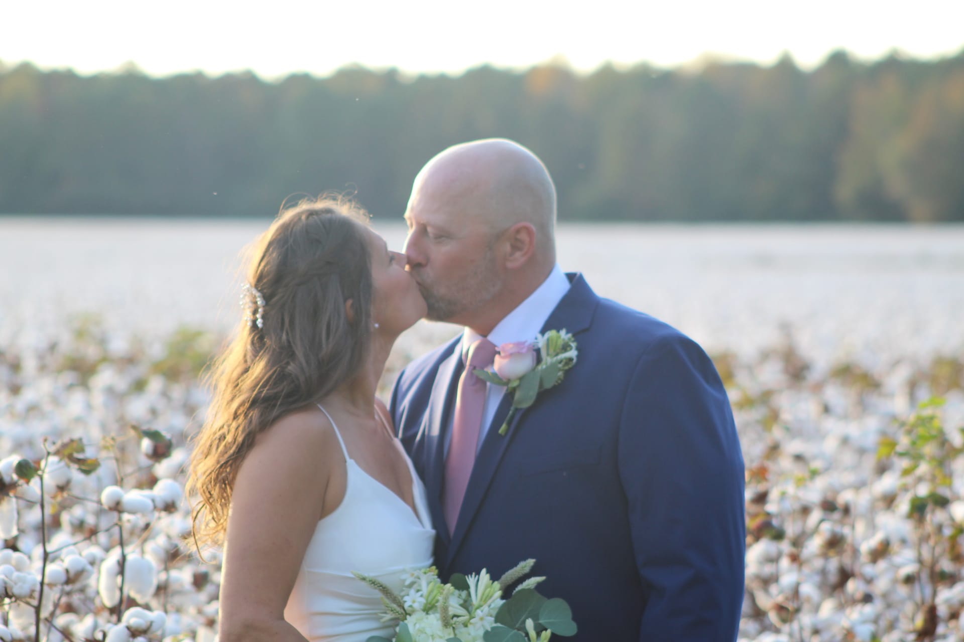 Bride and groom sharing a kiss at golden hour in a blooming cotton field with a tree line glowing in the background at Scarborough House.