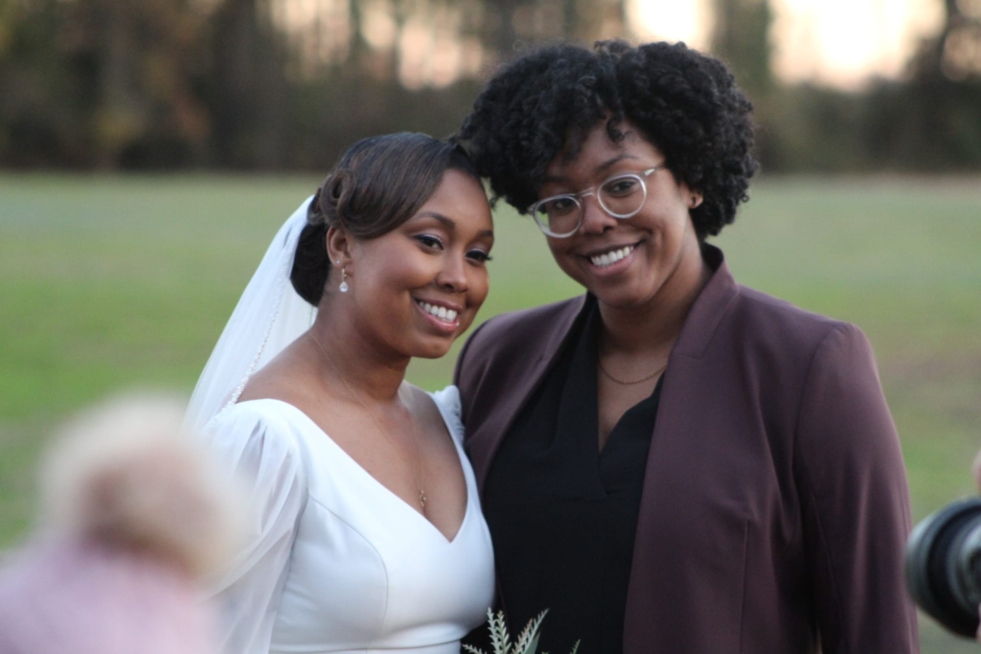 A smiling bride in a white gown and veil poses cheek-to-cheek with a guest in a burgundy blazer on the ceremony lawn at Scarborough House estate.